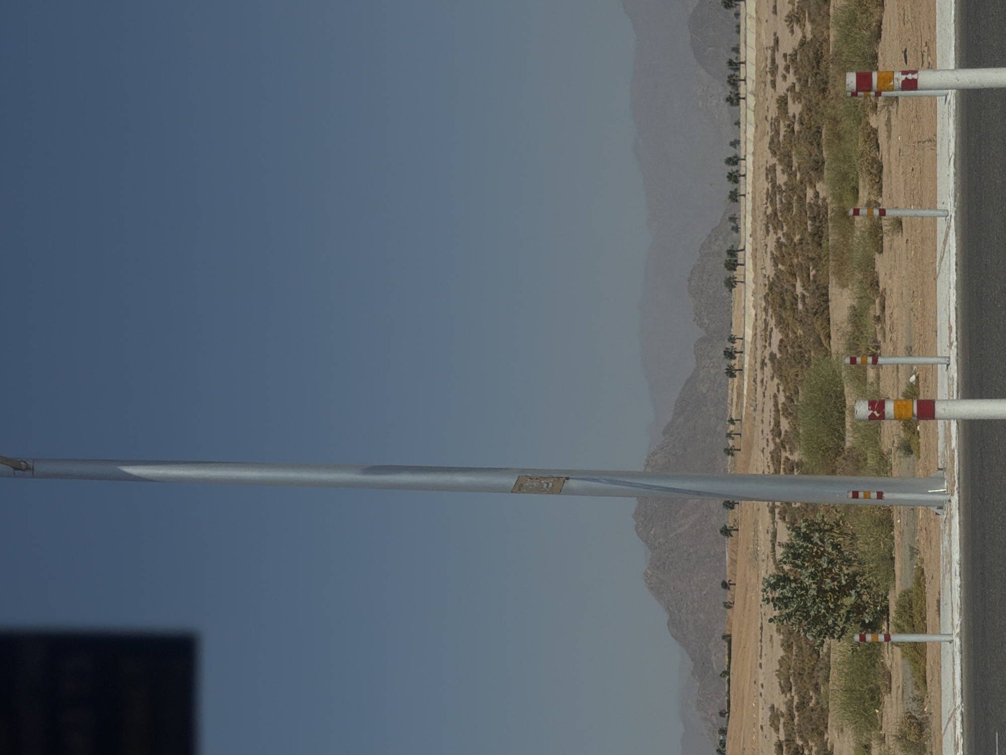 The image depicts a wide, clear blue sky above a desert landscape in Sharm El Sheikh, Egypt. In the foreground, a grey asphalt road is visible, bordered by a light grey curb and sidewalk. Several white bollards with red and yellow stripes are positioned along the sidewalk. To the left, a tall, thin, silver metallic pole stands prominently, with a small, rectangular emblem attached to it. In the middle ground, the desert terrain is characterized by sparse, dry vegetation and low bushes. A solitary, bushy green plant stands out amongst the dry scrub. Further in the background, a row of palm trees stretches across the horizon, silhouetted against a hazy mountain range. The mountains themselves are rocky and arid. The overall impression is one of a bright, sunny day in a desert environment, likely an arid region of Sharm El Sheikh.