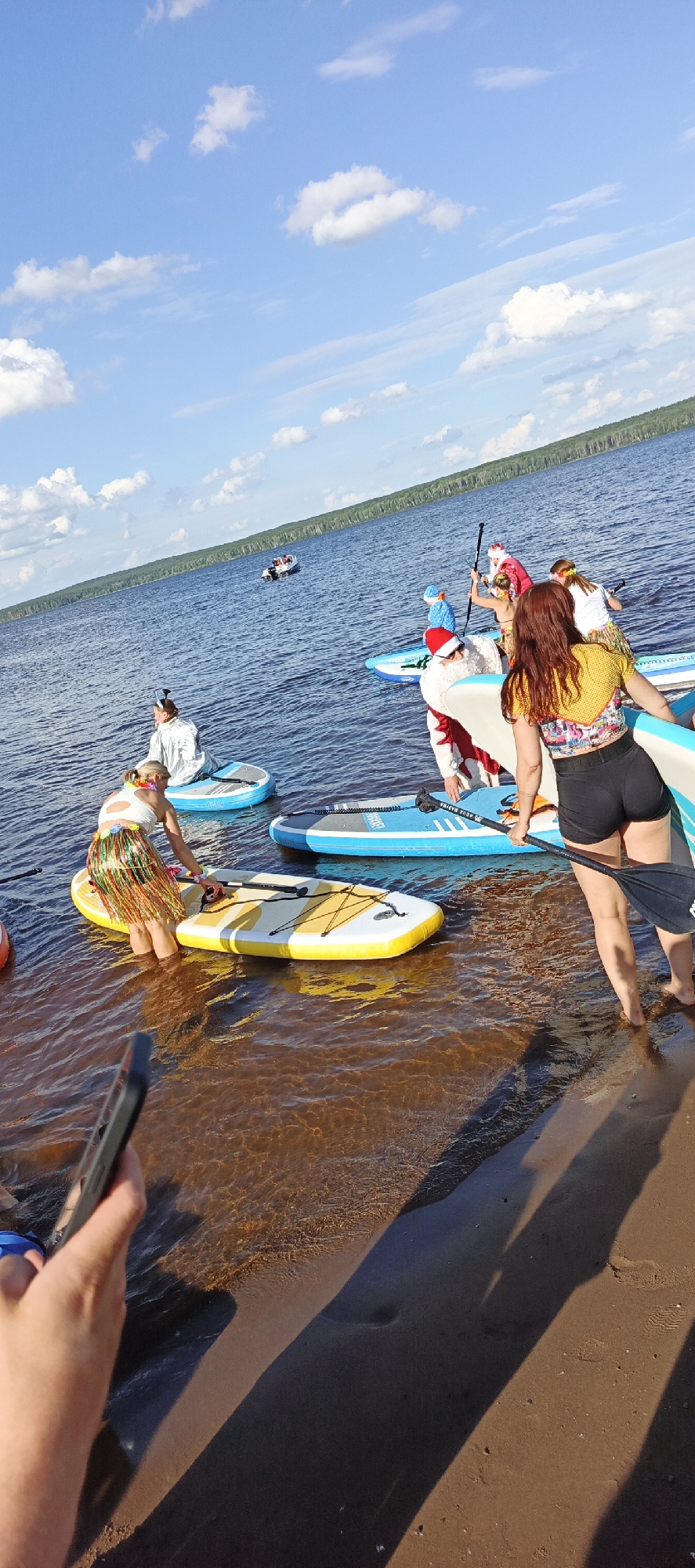 A group of people are paddleboarding on a lake on a sunny day. The water is calm and reflects the blue sky and white clouds. The background shows a dense forest lining the shore. Some individuals are dressed in Santa Claus costumes, indicating a festive event, possibly a holiday-themed paddleboarding gathering. One person is wearing a Hawaiian-style skirt. In the distance, a small boat with people in it is visible on the water. One person in the foreground is holding a phone, appearing to take a photo or video. The overall mood seems joyful and lighthearted. The visible text on one of the paddleboards reads "AQUA BAZINE". The scene suggests a warm, clear day, likely in the late morning or afternoon, given the shadows cast. The location context provided is Chaikovsky, Russia.