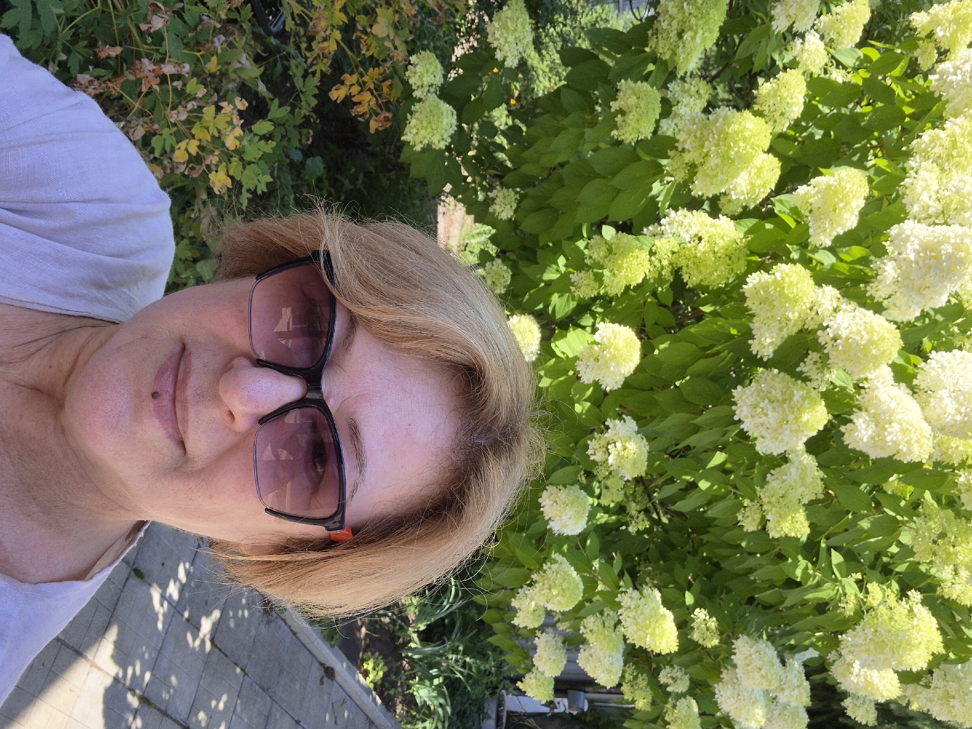 A woman with blonde hair and glasses is taking a selfie outdoors. She is wearing a light-colored top. Behind her is a large bush with clusters of white and yellow flowers and green leaves. The background also shows a paved walkway and some other greenery. The scene is set in the city of Malaya Tsarevschina, Russia.