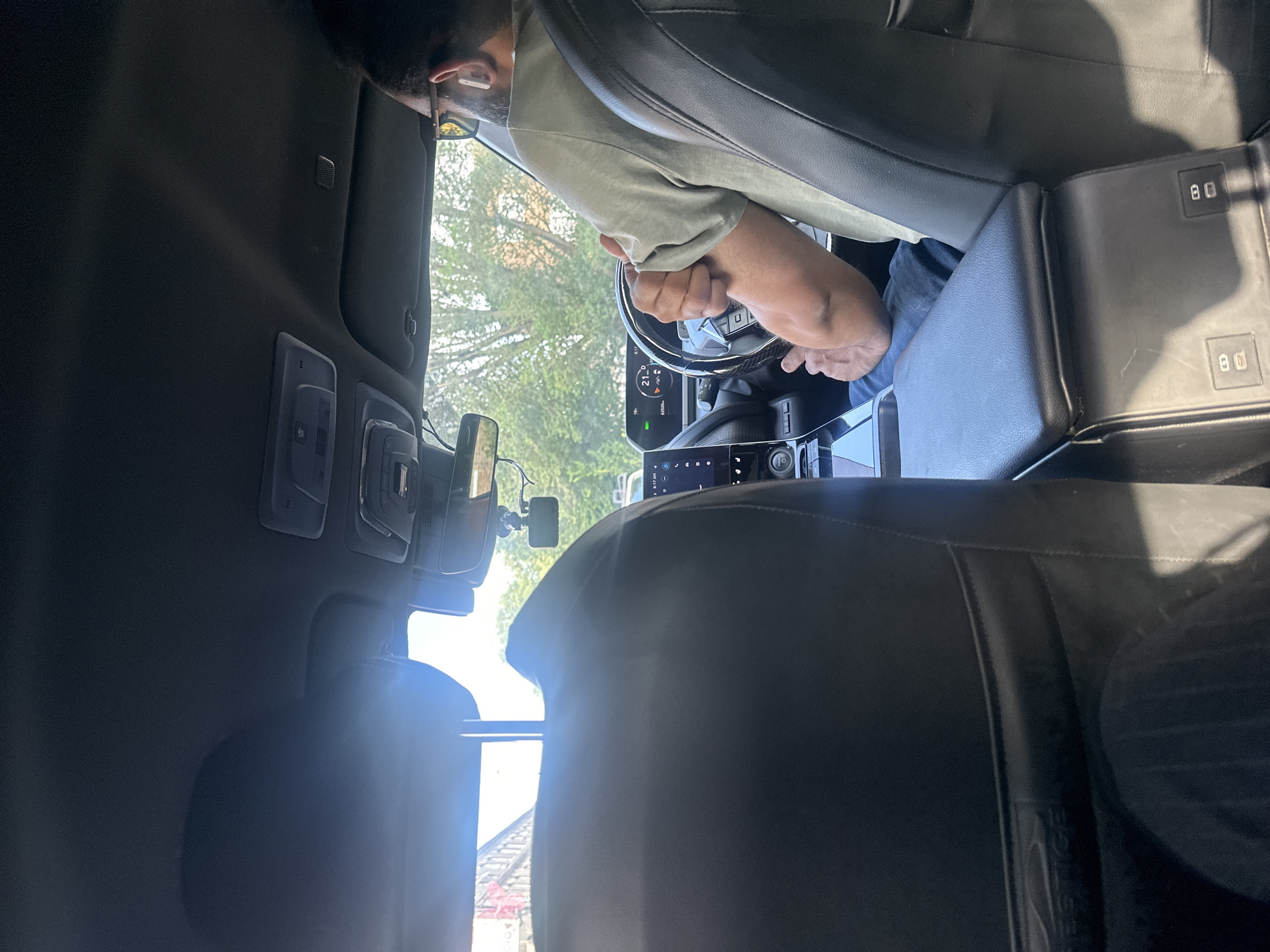 The image shows the interior of a car, likely in London, United Kingdom. A person with sunglasses and AirPods is driving. They are wearing a light green t-shirt and blue jeans. The car's dashboard shows a temperature of 21 degrees Celsius and a speed of 21 mph. The sun is shining, and trees are visible outside the car windows, suggesting it is daytime. The interior of the car features black leather seats and a center console. There is a dashcam mounted on the windshield.