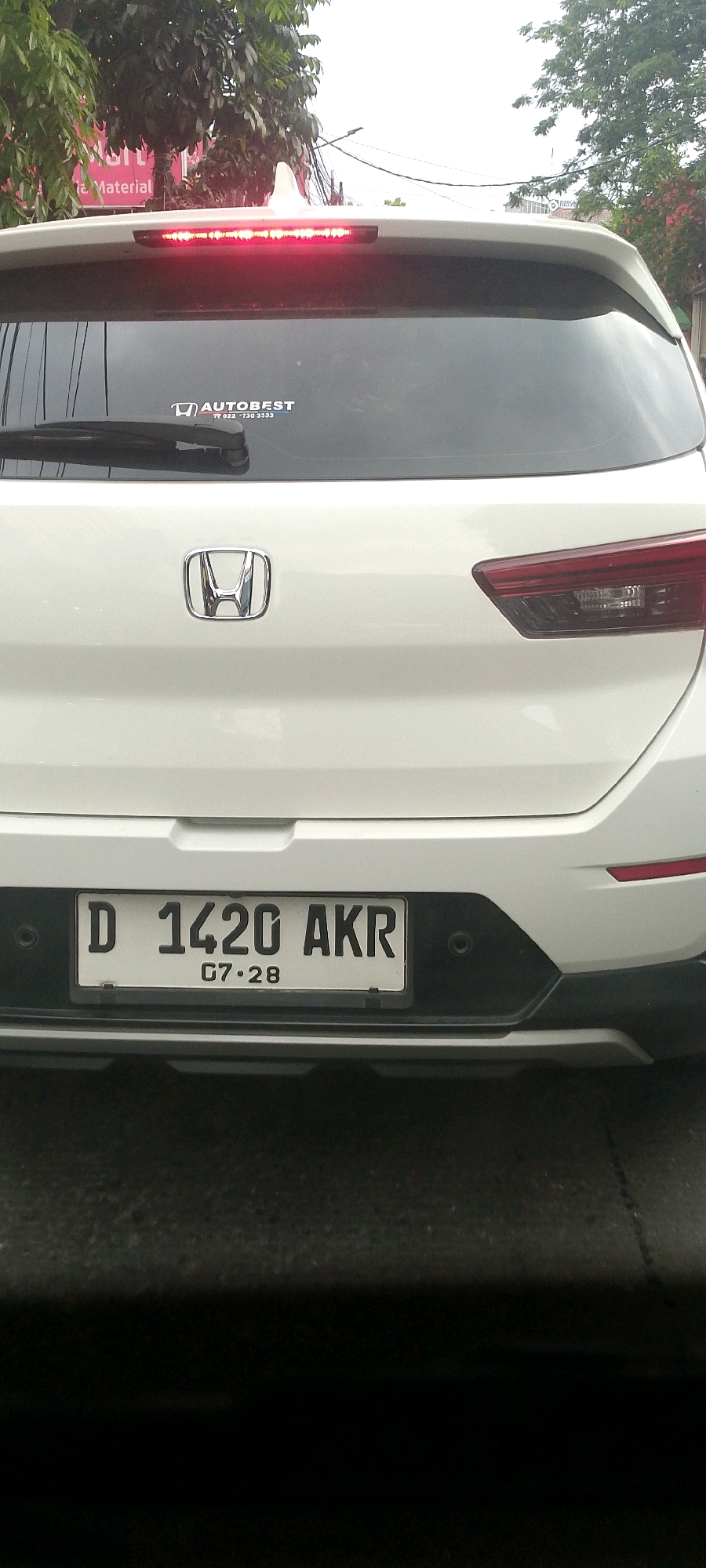 The image shows the rear of a white Honda car, likely in Bandung City, Indonesia, as indicated by the license plate format. The license plate reads "D 1420 AKR" and has the expiry date "07·28" below it. A red brake light is illuminated across the top of the rear window. A sticker with the text "AUTOBEST" and a phone number is visible on the rear windshield. The car has a Honda logo centered on the trunk. The background features trees and some buildings, suggesting an urban or suburban environment. The image is captured from a slightly low angle, looking upwards at the rear of the vehicle.