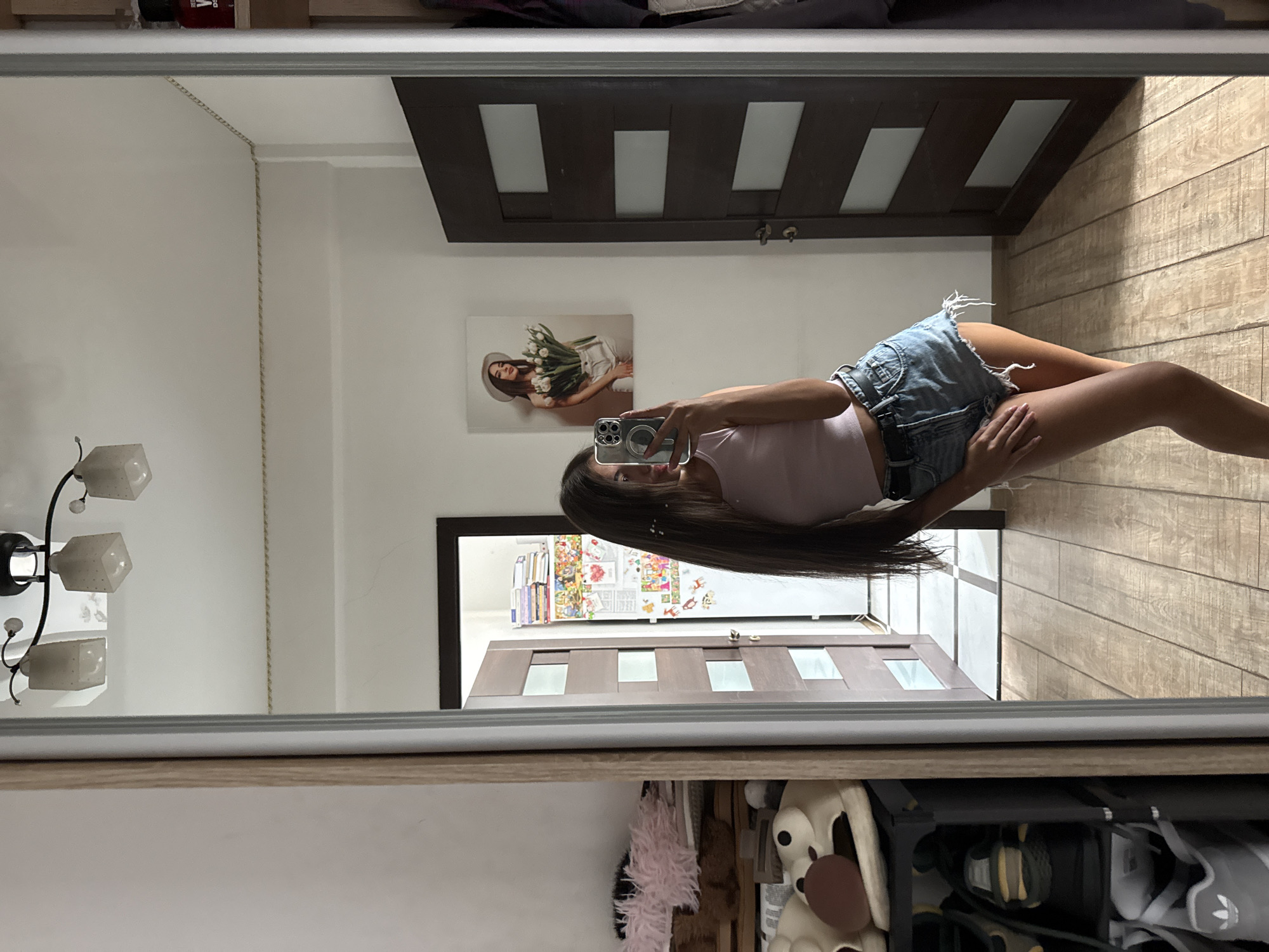 The image captures a young woman taking a mirror selfie in what appears to be a residential setting. She is standing in a hallway, with a large mirror reflecting her image. She is wearing a pink tank top and denim shorts. In the reflection, we see a door with frosted glass panels, a picture of a woman holding flowers on the wall, and a shelf with books and a red bottle. To the left of the mirror, there's a glimpse of a shoe rack with several pairs of shoes and some fluffy pink items on a shelf. The floor is made of wooden planks. The environment suggests an indoor domestic space.
