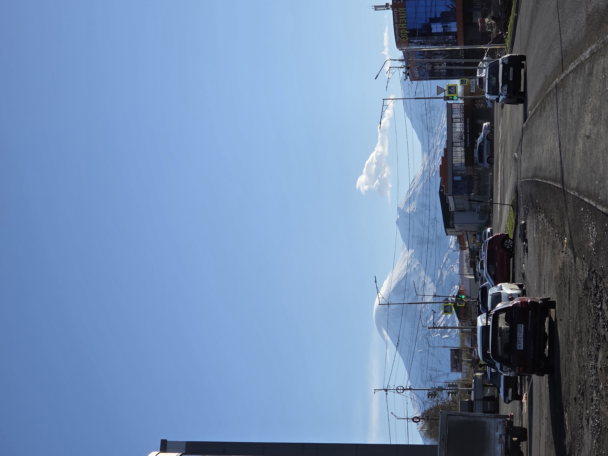 The image depicts a street in the city of Unknown, Unknown, with snow-capped mountains in the background. The sky is clear and blue. Several cars are parked along the right side of the street. Buildings and power lines are visible along the street. A sign with Cyrillic characters is prominently displayed on a building.
