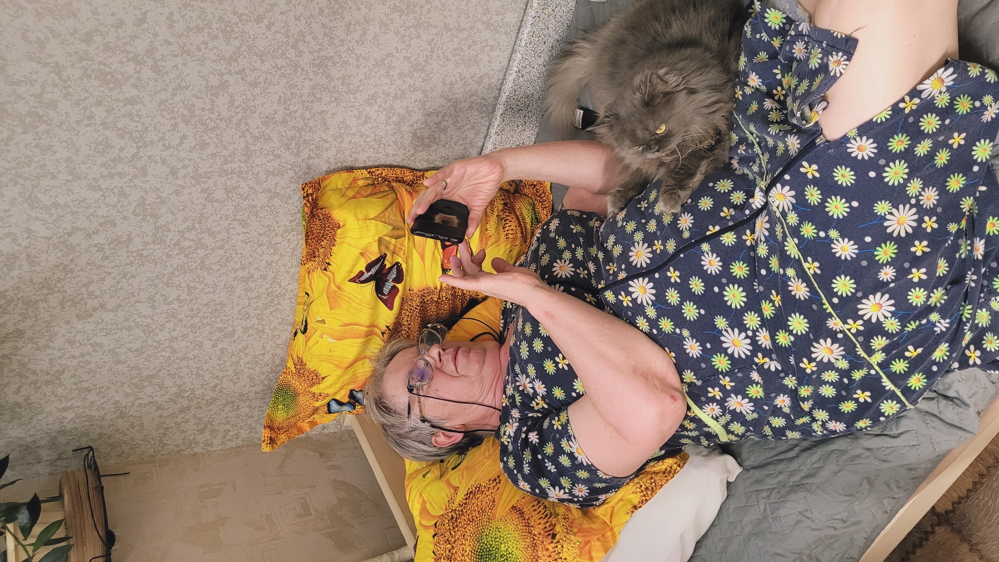 An elderly Caucasian woman with grey hair, wearing glasses and a dark blue dress with a floral pattern, is lying on a bed, looking at her smartphone. Beside her, a long-haired grey cat with yellow eyes is also on the bed. The bed is covered with a grey sheet and a yellow pillowcase with a sunflower and butterfly print. The background is a textured beige wallpaper. The scene appears to be indoors.
