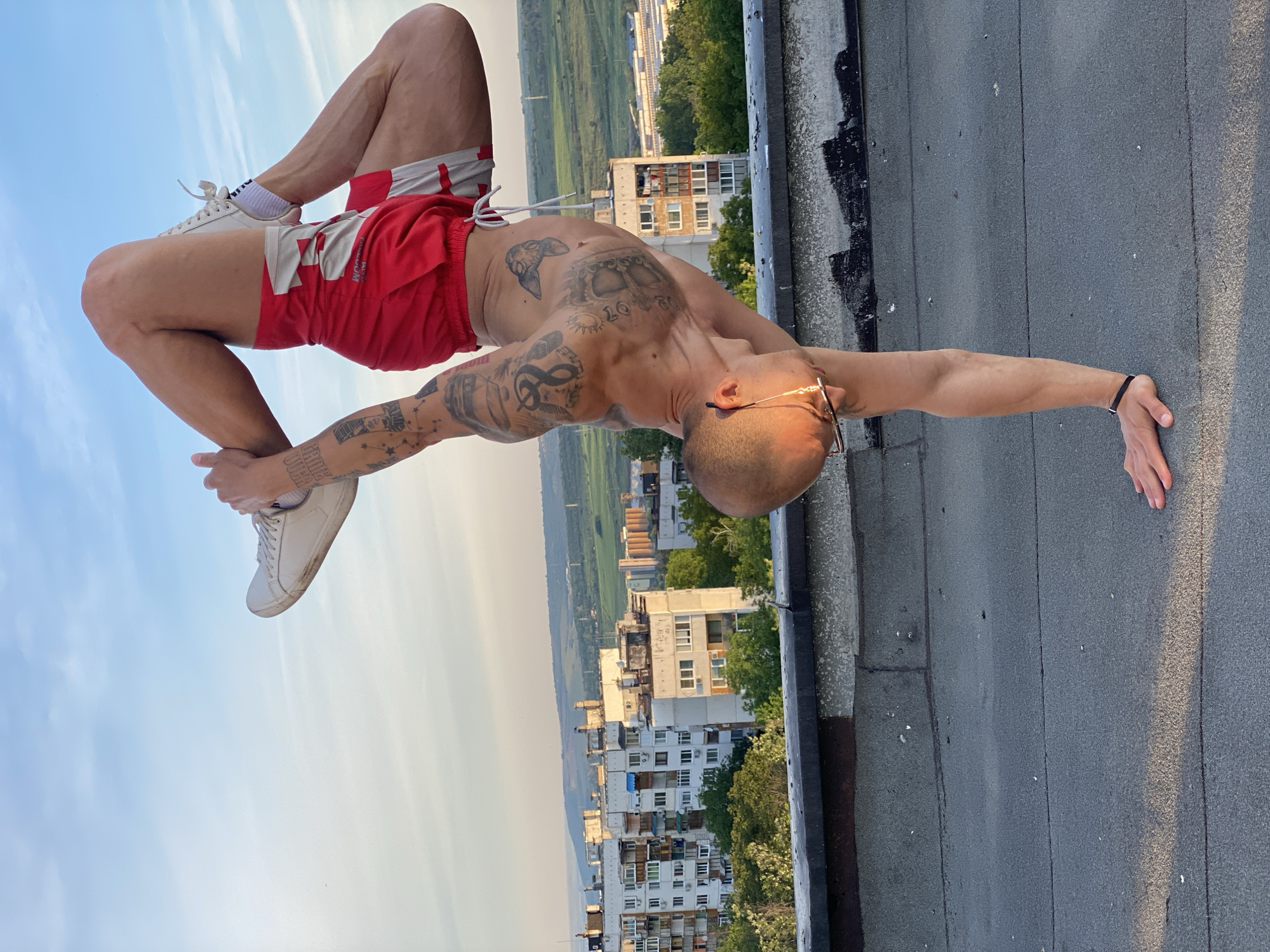A man is performing a contortionist pose on a rooftop, with the city of Pleven, Bulgaria, in the background. The man is bald, has multiple tattoos on his torso and arms, wears glasses, a black wristband, red and white shorts, white shoes, and white socks. He is bending backward, resting his hand on the roof and placing his feet near his head. The rooftop is gray with black edging, and the cityscape beyond includes buildings, trees, and a distant horizon under a hazy sky.