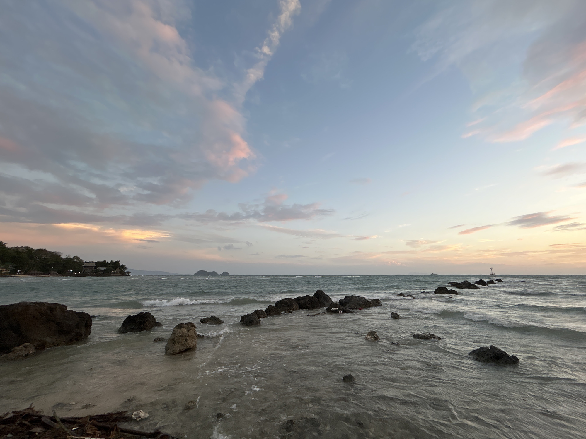 The image depicts a serene coastal scene during what appears to be sunset or dusk in Ko Pha-ngan District, Thailand. The sky is a soft gradient of blue, pink, and orange, with wispy clouds scattered across it. A calm sea with gentle waves stretches towards the horizon, where several small islands are visible in the distance. Rocks of various sizes dot the shoreline and protrude from the water, with some forming a partial breakwater. On the left side of the image, lush green trees and buildings suggest a populated area along the coast. The foreground consists of a sandy beach with scattered debris, including driftwood. The overall atmosphere is tranquil and picturesque, characteristic of a tropical beach at the end of the day.