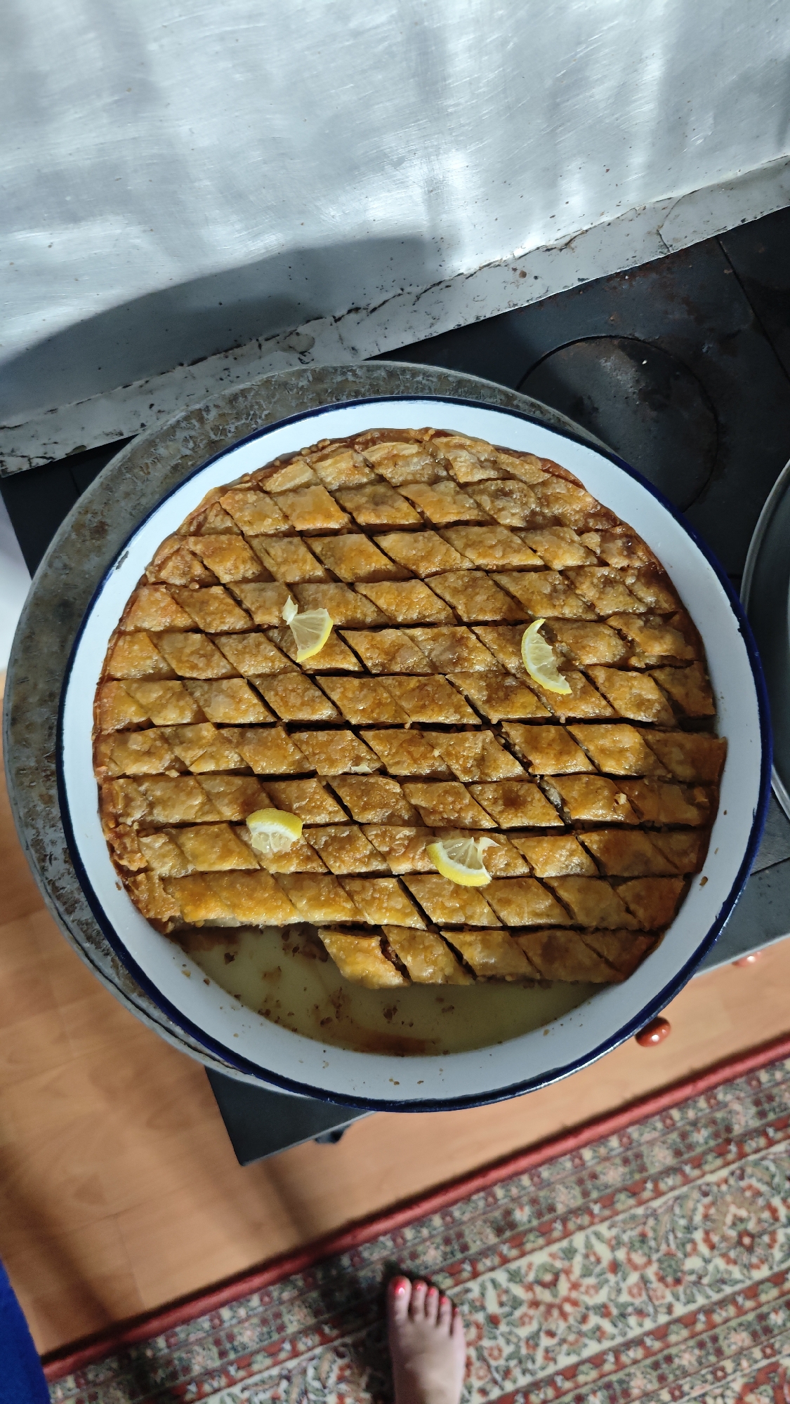A top-down shot shows a large, round pan of baklava, garnished with lemon slices. The baklava is cut into diamond shapes and has a golden-brown color. The pan has a white interior and a blue rim. The pan is sitting on a surface made of metal with signs of wear. To the left, a white surface is visible. Below the pan, there's a glimpse of a wooden floor and a section of a patterned rug, with a foot near the edge of the frame. The location is Planinitsa, Bulgaria.