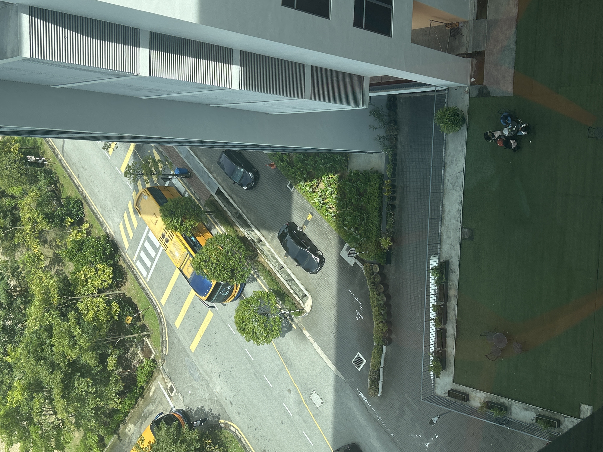 The image is a high-angle view, possibly from an upper story of a building, overlooking a street scene. The scene includes multiple vehicles, including a yellow and blue taxi, as well as black cars parked along the side of the road. There is a building with grey facade, including venetian blinds. Some individuals are seated at a round table on a grassy area. The setting appears to be Kuala Lumpur, Malaysia. The road has yellow pedestrian crossing markings. The environment includes greenery with shrubs and trees.