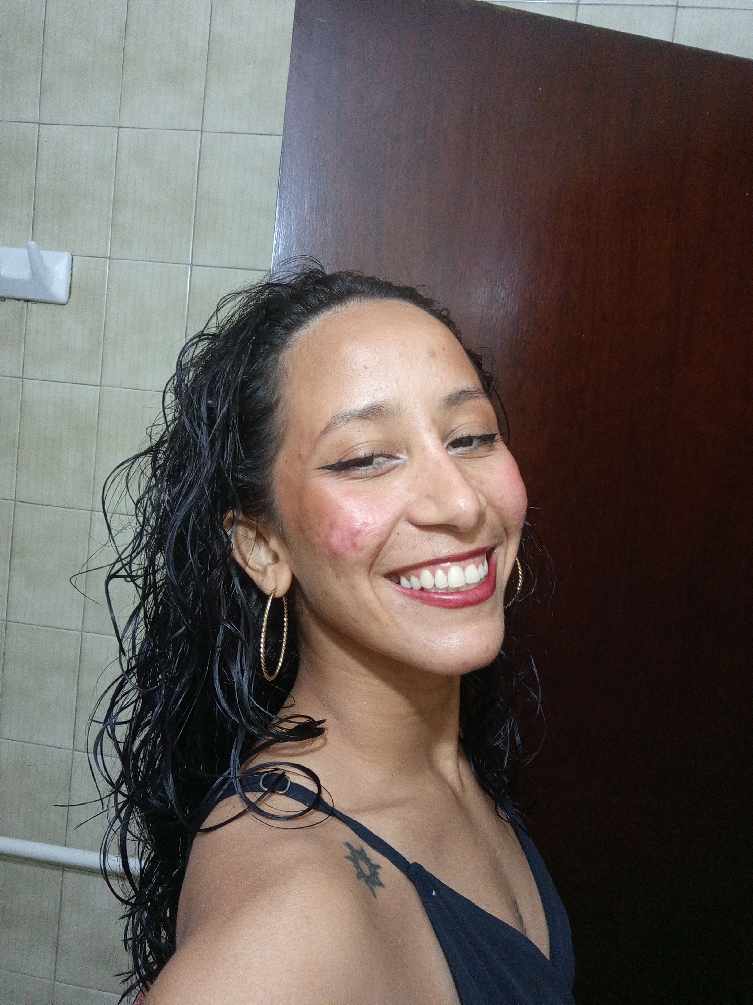 A woman with wet, dark curly hair smiles at the camera. She wears gold hoop earrings and a black tank top. A tattoo is visible on her left shoulder. Behind her are a tiled wall and a dark brown door or panel. Based on the location tag, this image was taken in Porlamar, Venezuela.