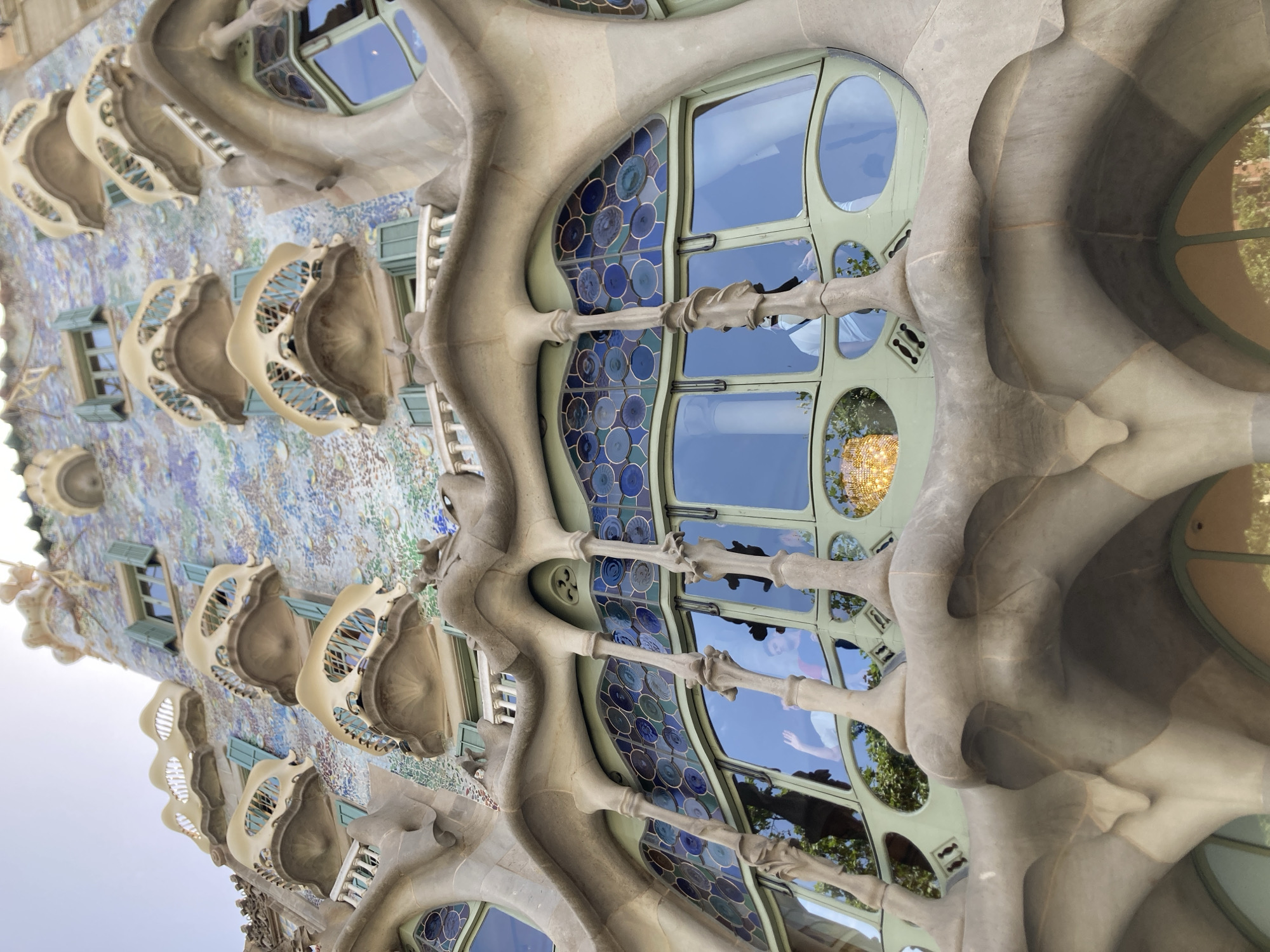 The image depicts a close-up, low-angle view of the facade of Casa Batlló, a renowned architectural masterpiece designed by Antoni Gaudí, located in Barcelona, Spain. The building's exterior is characterized by its undulating, organic forms, reminiscent of bone structures and nature. The facade is adorned with a mosaic of colorful ceramic tiles, predominantly in shades of blue, green, and purple, creating a shimmering, iridescent effect.
The central focus of the image is a series of large, arched windows framed by what appear to be sculpted stone balconies. These balconies have a fluid, bone-like appearance and are decorated with intricate detailing. The windows themselves are segmented into smaller, circular panes of stained glass, featuring concentric circles of various shades of blue. The reflections in the windows reveal glimpses of trees and the sky, as well as at least two individuals inside the building. One individual, wearing a light-colored shirt and a face mask, is visible waving.
Above the main window structure, several smaller windows with light blue shutters are visible, set against the mosaic-covered wall. The overall impression is one of whimsical, almost surreal design, showcasing Gaudí's distinctive Art Nouveau style. The lighting suggests it is daytime, with the sun casting a warm glow on parts of the building and reflecting in the windows. The perspective emphasizes the height and intricate details of the architecture.