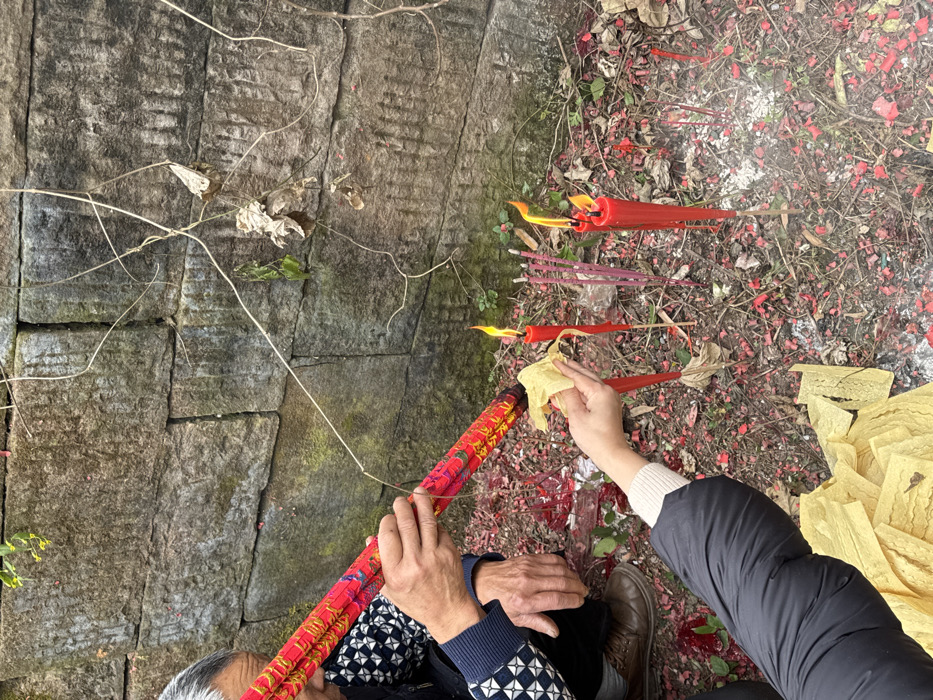 Two people are pictured from an overhead perspective, possibly performing a ritual. One person holds firecrackers, and another holds a yellow cloth near burning red candles and incense sticks amidst scattered red debris on the ground. A stone wall is on the left. The scene suggests a traditional ceremony or offering.