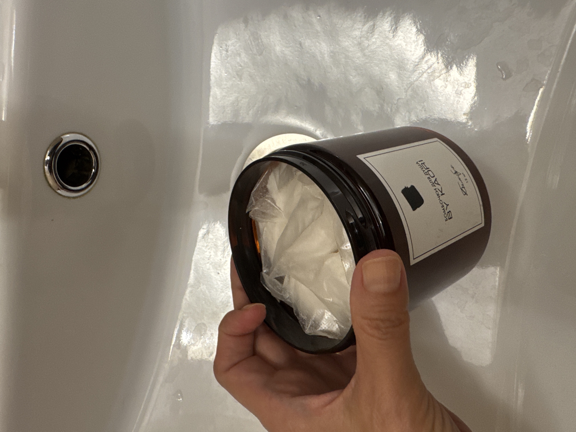 The image shows a hand holding a brown glass jar with a white label, containing several small, individually wrapped white bath bombs. The jar is positioned over a white ceramic sink with a chrome drain. The sink appears to be wet with some water droplets visible. The label on the jar is partially obscured by the hand and the contents, but text including "БОМБОЧКИ ДЛЯ ДУША BY KAORI" and a signature can be seen. The background is a close-up of the sink's surface and the drain. There are no clear indicators of the specific location within Saint Petersburg.