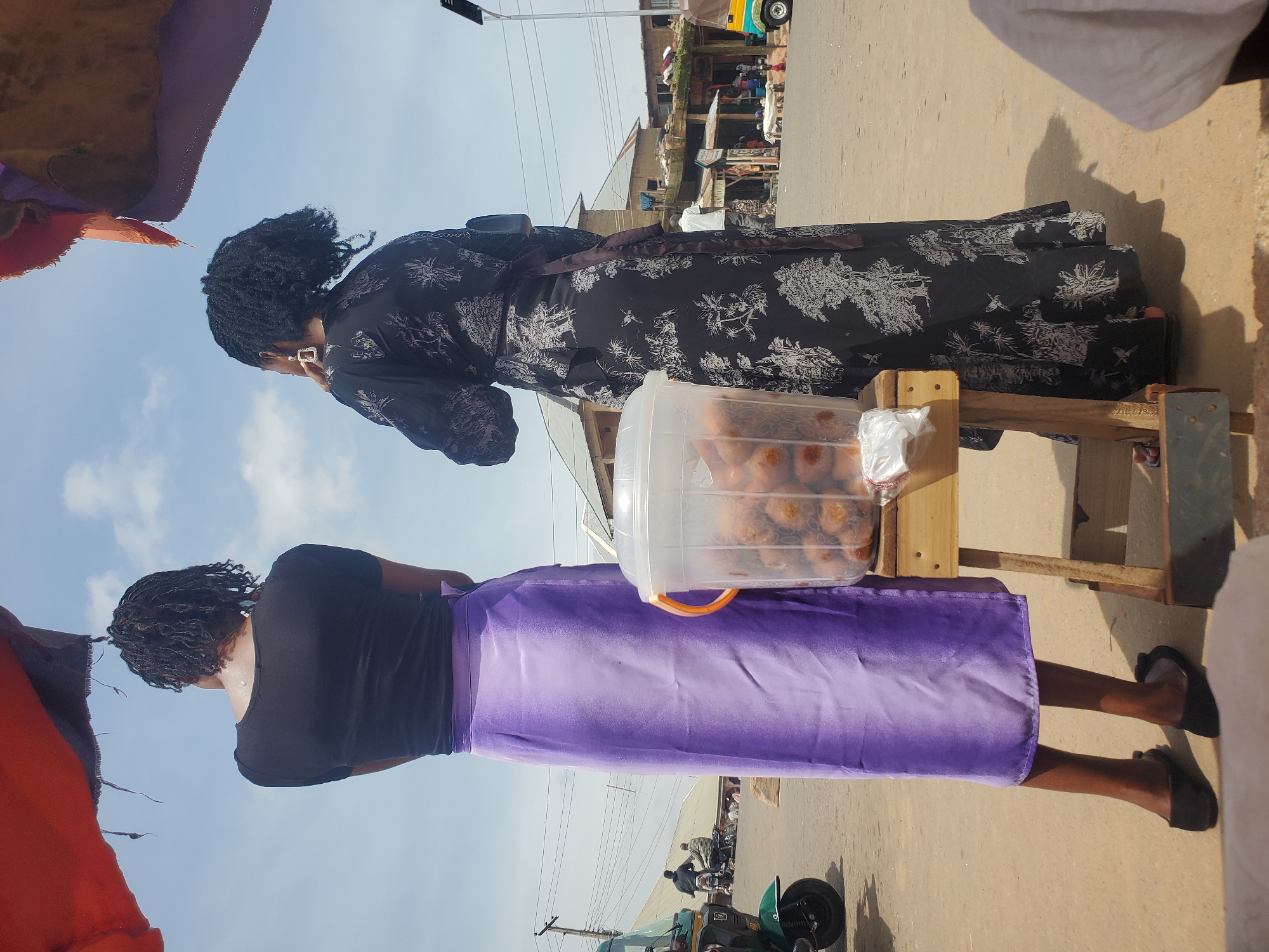 The image depicts two women standing in what appears to be a bustling outdoor market in Gwong, Nigeria. The overall lighting suggests it is daytime, possibly mid-morning or afternoon, with clear skies.
The woman in the foreground is wearing a black short-sleeved shirt and a long, flowing purple skirt. She has braided hair and is looking away from the camera, her stance suggesting she might be a vendor or a shopper. To her left, a large clear plastic container filled with what looks like baked goods or snacks is visible on a wooden stand. Next to the container is a white plastic bag.
Behind her, another woman is also facing away from the camera. She is wearing a long, black dress with a white intricate pattern. Her hair is styled in braids, and she appears to be wearing earrings. She is standing near the same wooden stand.
The background shows a busy street scene with buildings, power lines, and other market stalls. A yellow and blue motorized tricycle, often used for transportation or carrying goods in Nigeria, is visible in the distance, along with other people and vehicles. The ground is paved, and there is a general sense of commercial activity. The clothing styles and the presence of the tricycle are strong indicators of the location in Nigeria.