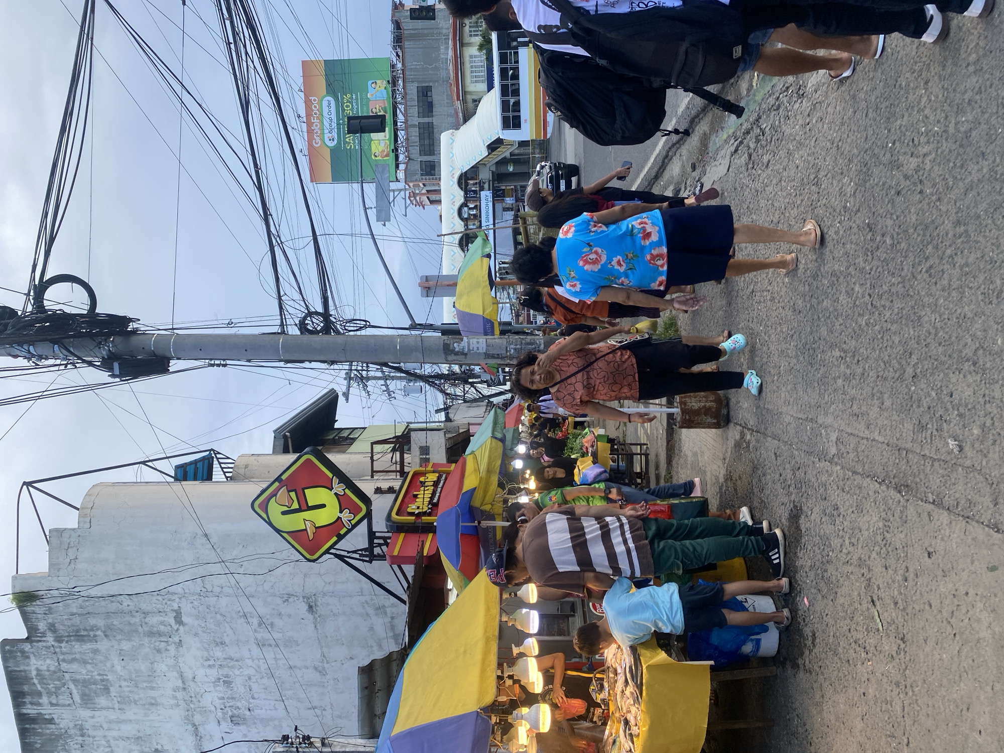 The image depicts a bustling street scene in Davao City, Philippines, likely during daylight hours. A street market is in full swing, with vendors and customers gathered around stalls adorned with colorful umbrellas and illuminated by hanging lights. Several people are seen standing and walking along the paved street, some looking at goods displayed on tables. The overall atmosphere suggests a lively commercial area with a mix of shoppers and people going about their day.
Prominent in the image is a stall selling fish, with rows of fresh catch laid out on a yellow table. A young boy in a blue shirt is seen inspecting the fish, while other individuals, possibly vendors, are also present at the stall. Nearby, another vendor is attending to a customer at a stall with a variety of items. The market is set against a backdrop of buildings and a prominent billboard advertising "GrabFood" with the tagline "Group Order. Save Together. 30% Off."
Above the market, a tangle of electrical wires crisscrosses the sky, a common sight in many urban areas in the Philippines. A traffic light is also visible, suggesting the proximity of an intersection. In the background, a bus and other vehicles are present, indicating ongoing traffic. A large, somewhat weathered building with a distinctive diamond-shaped sign featuring a yellow character is also visible. The scene is crowded and dynamic, conveying a sense of everyday life and commerce in a Philippine city. The weather appears overcast, contributing to a soft, diffused light across the scene.