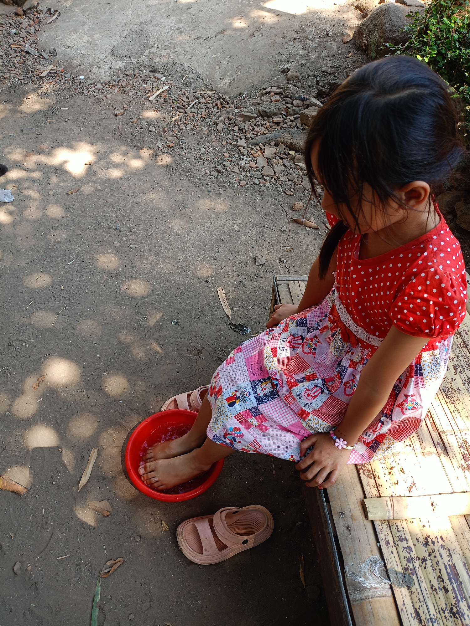 A young girl with dark hair, wearing a red and white polka dot top and a patchwork dress, sits on a bamboo bench. Her feet are submerged in a red basin filled with water. A pair of light brown sandals lies on the ground beside the basin. The ground around her is a mixture of dirt, gravel, and scattered leaves. In the background, there are large rocks and green foliage. The scene appears to be outdoors, possibly in a rural area of Jelbuk, Indonesia, with sunlight casting dappled shadows on the ground.