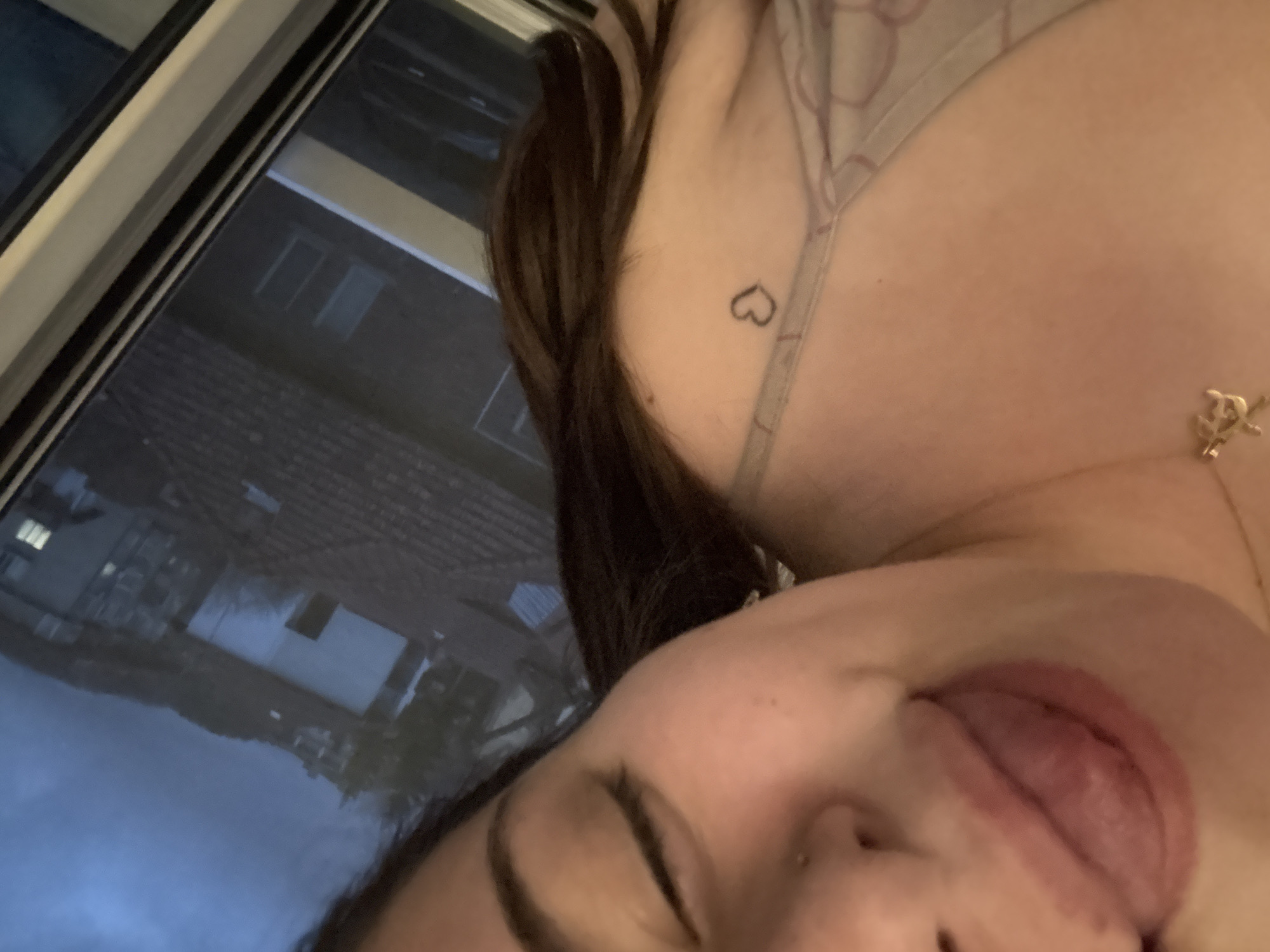 The image depicts a close-up, upward-angled view of a female individual indoors. Her face is shown from the nose down, with her eyes closed or heavily downcast, and her tongue protruded from her mouth. A small stud piercing is visible on her left nostril. She wears a light-toned, patterned spaghetti-strap garment. A gold-toned necklace with a stylized "CC" pendant rests around her neck, and a small, dark heart tattoo is located on her left shoulder. Dark hair cascades over her right shoulder.
The background, viewed through a reflective window, presents an exterior cityscape identified as Troyan, Bulgaria. The sky is dark grey, suggesting low ambient light, such as dusk or an overcast day. Dark-tiled rooftops populate the immediate exterior view. A prominent white building with a dark, rectangular opening, suggestive of an entrance or garage door, is visible. Beyond, residential structures with illuminated windows are discernible on a hillside. A dark, prominent hill or mountain range forms the distant horizon, interspersed with sparse vegetation. A multi-pane window frame, composed of white and dark elements, borders the right side of the image.