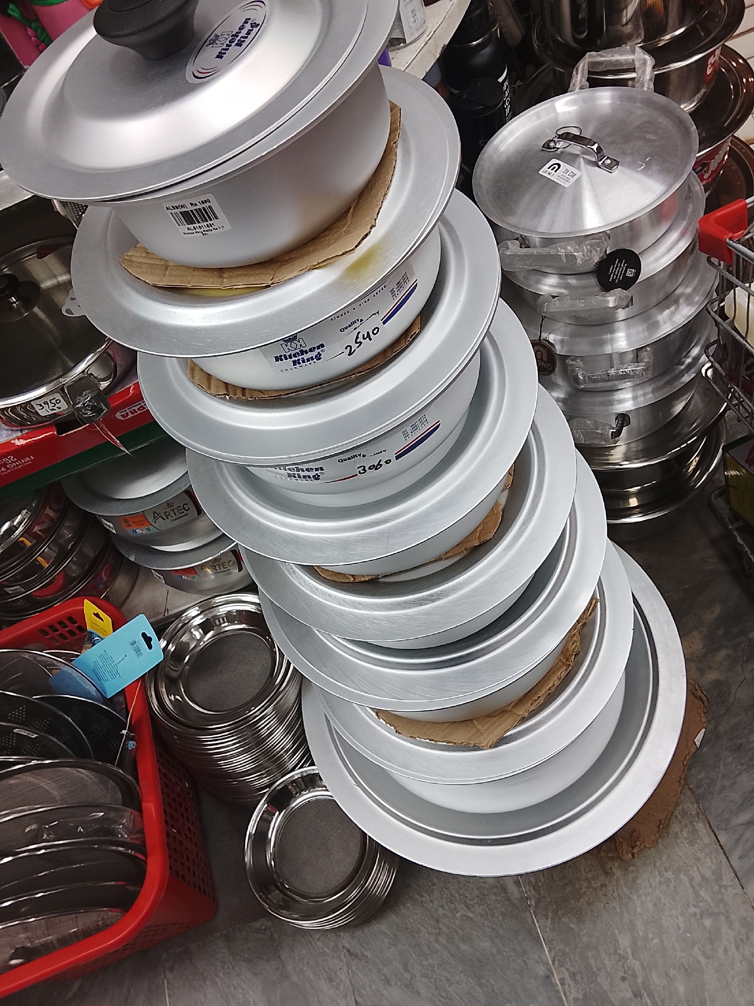This is a still, high-angle shot depicting a display of aluminum cookware and kitchenware at a shop, possibly in Gulgasht Colony, Pakistan. Dominating the scene is a precarious stack of "Kitchen King" brand pots and pans, separated by pieces of cardboard. To the left, a red plastic basket holds an assortment of strainers and other metallic kitchen tools. In the background, there are additional pots and pans, including some marked "ARTEC". The floor is tiled in a dark gray pattern. Other objects like a bottle and a shopping cart are partially visible in the background. There are labels showing prices on some of the items, such as "Rs 1600" and "2540".