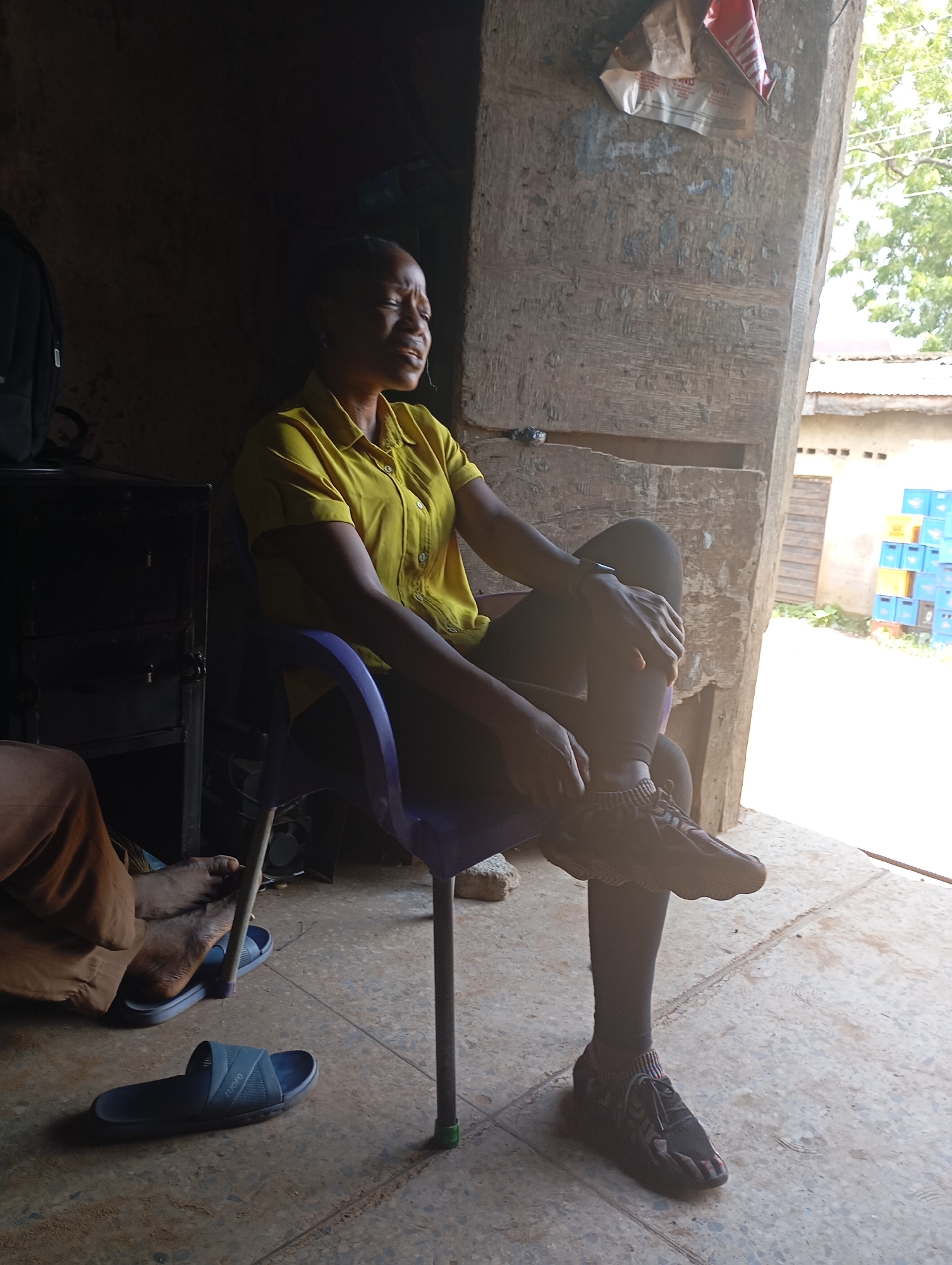 A woman sits in a purple plastic chair in what appears to be an outdoor or semi-enclosed space. She is wearing a yellow collared shirt and black leggings, with dark athletic shoes on her feet. Her left hand rests on her left knee, and her right arm is bent, with her hand near her face. Her eyes are closed, and her mouth is open, suggesting she may be speaking, singing, or in distress. To her left, a dark cabinet or shelf unit is visible. In the foreground, on the concrete floor, are two pairs of dark blue sandals. Behind her, a rustic wooden structure is prominent, with a piece of torn packaging attached to it. Sunlight streams in from the right, illuminating the scene and casting shadows. In the background, a stack of blue and yellow crates and trees are visible, indicating an outdoor or open-air environment within the city of Shendam, Nigeria.