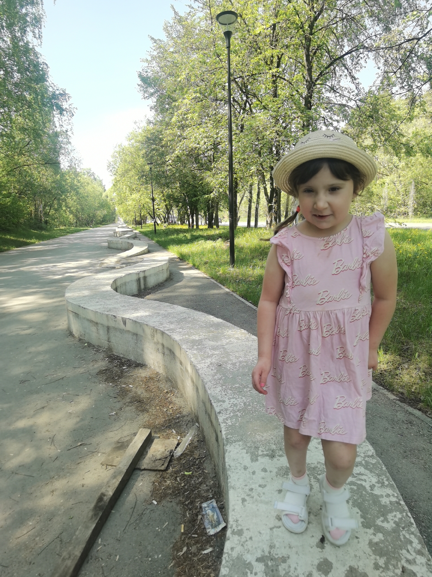 The photo depicts a young girl standing on a concrete structure in what appears to be a park or recreational area. The girl is wearing a pink dress with "Barbie" printed on it and a light-colored hat with a dark band. She is wearing white sandals with socks. The structure she is standing on is a wavy, low concrete wall. Behind the girl is a paved pathway lined with trees, with a lamp post visible in the background. The photograph was taken in the city of Perm, Russia.
