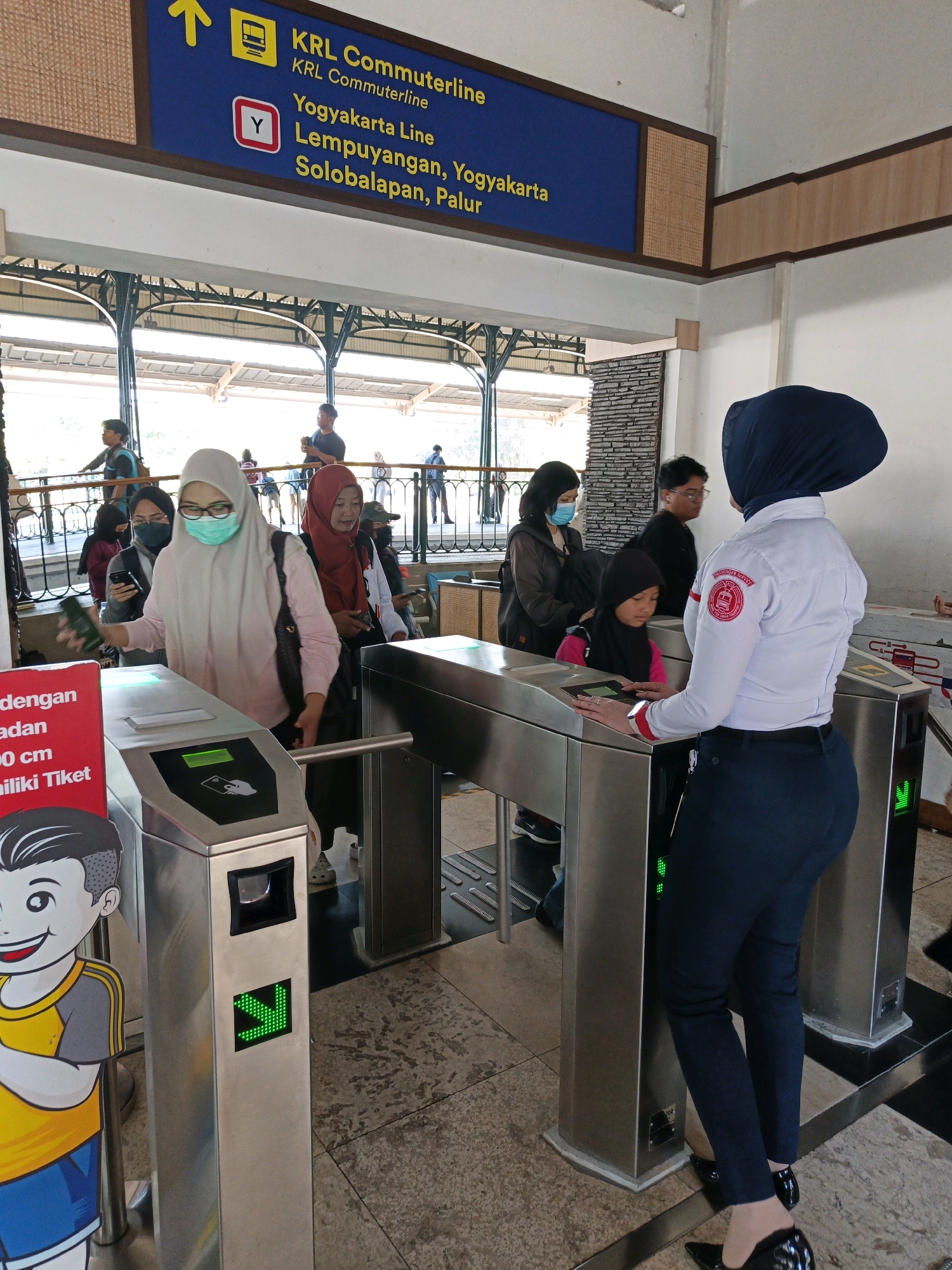 An indoor view of a train station ticket gate in Klaten, Indonesia, shows several individuals interacting with the automated barriers. A woman wearing a navy hijab and a white uniform shirt with an emblem that reads "KAI ACCESS" stands to the right, facing away from the camera and towards the ticket gates. She appears to be an attendant, with her right hand resting on a ticket gate. In front of her, a woman wearing a light pink hijab and a light blue medical mask is passing through a ticket gate, holding a smartphone in her left hand. Another woman wearing a red hijab is to her left, and behind them, several other people, including a child, are visible. The ticket gates are made of stainless steel, with green arrow indicators on the front. A red sign with white text is attached to the left-most ticket gate, and a cartoon image of a boy is to the left of that. Above the ticket gates, a blue sign with yellow text indicates train routes, including "KRL Commuterline" and destinations for "Yogyakarta Line". The environment appears to be a covered walkway with architectural elements suggesting a public transport hub.