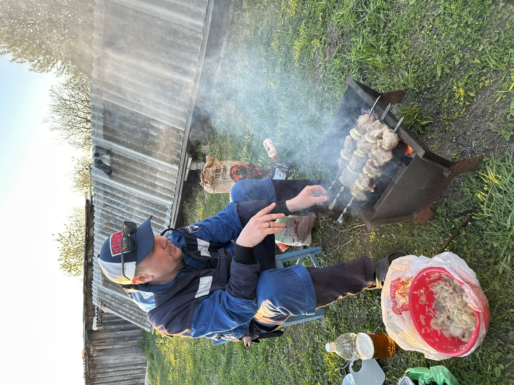 A man is sitting on a small blue folding chair, tending a barbecue grill with skewers of meat and onions. He is wearing a blue jacket, dark pants, and a blue baseball cap with sunglasses perched on the brim. The grill is emitting smoke. Next to the man is a red bowl filled with what appears to be marinated meat, along with a water bottle and other items. The setting is an outdoor area with a shed with corrugated metal siding in the background. The location is in the city of Среднее Кечево, Russia.