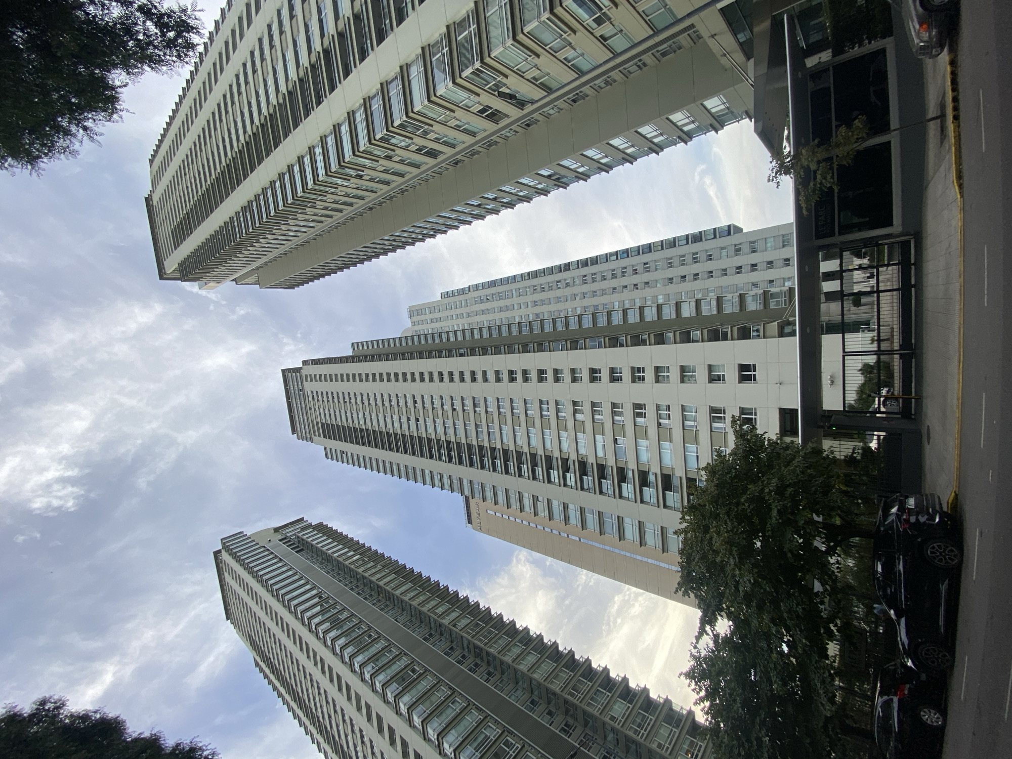 Low-angle view of tall residential buildings in the city of Buenos Aires, Argentina. Three main buildings, constructed with modern architectural design with repetitive window patterns, stretch high into the sky. Buildings appear as if they are reaching towards the cloudy sky. The building complex on the right has a sign reading "LE PARC" above the name "FIGUEROA ALCORTA". Street view on the right, including parked cars and a street edge. Some trees present at the bottom and edges of the frame. The image suggests an urban setting with high-rise apartment complexes.