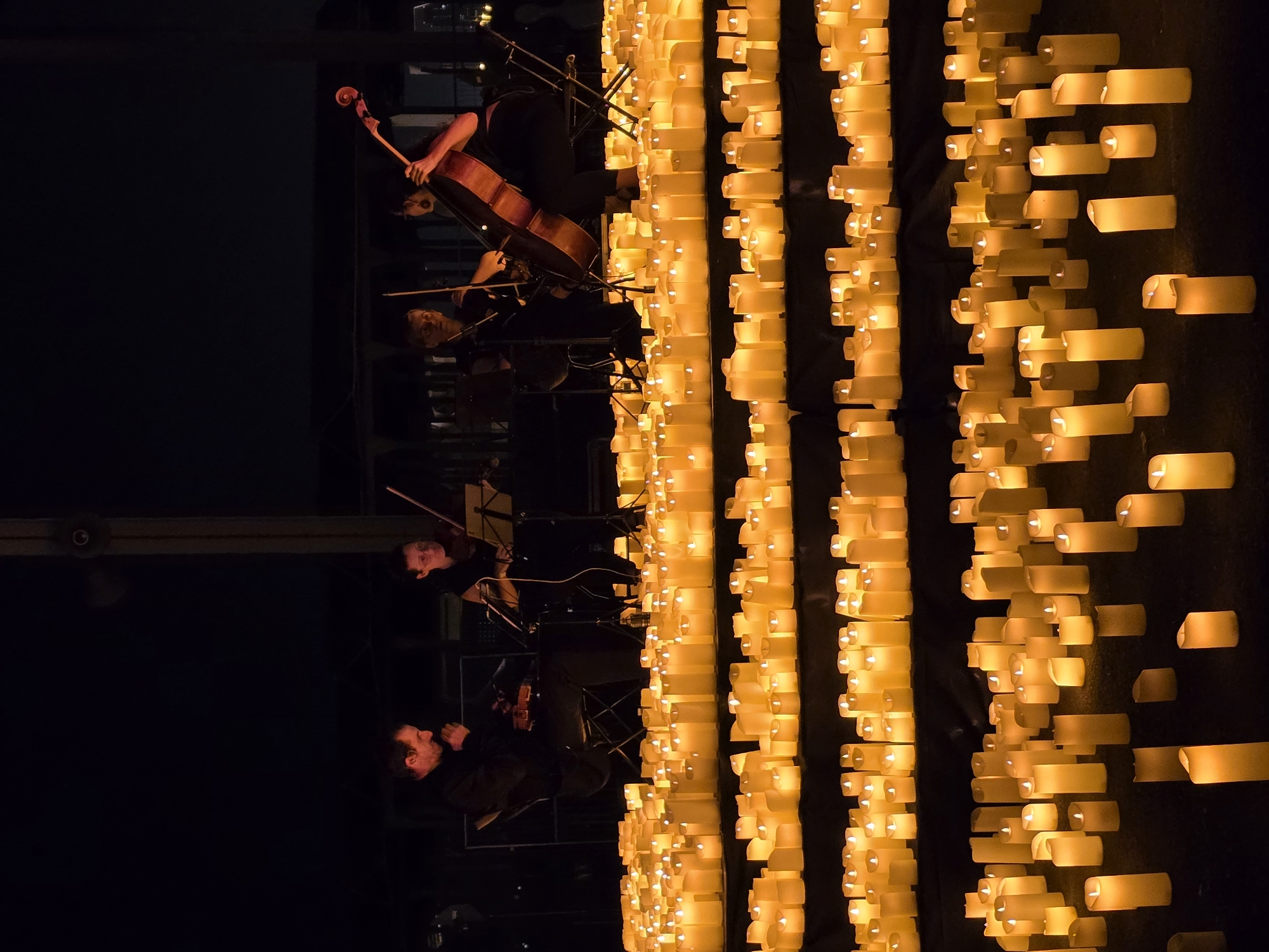 The image shows an outdoor nighttime scene at the Parc del Fòrum in Sant Adrià de Besòs, Spain, featuring a string quartet performing in front of a multitude of lit candles. There are four musicians on a raised platform, two on the left and two on the right. The musician on the far left is playing a violin, while the musician to their right is also playing a violin. The musician on the far right is playing a cello. The fourth musician, to the left of the cello player, is playing a viola. They are all seated and appear to be engrossed in their performance. The stage and surrounding area are covered in rows of white LED candles, creating a warm, ambient glow. The background is dark, with some indistinct structures visible, suggesting an open-air venue. The overall atmosphere is somber and serene.