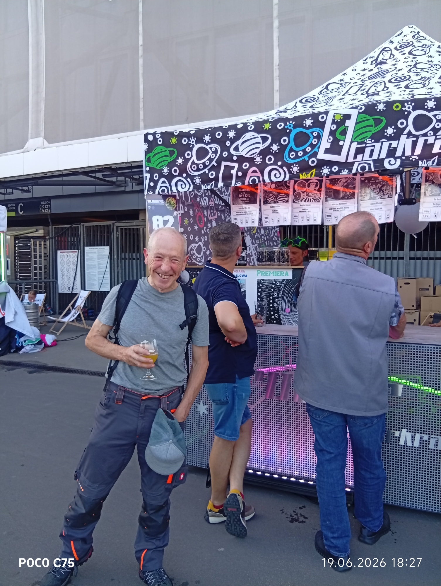 A man in a grey t-shirt, black backpack, and grey work pants with orange accents smiles and holds a glass of beer while standing outside a tent. The tent has a black and white pattern with alien and planet designs. Behind him, a bar area is set up with menus displaying drinks. Two other men are visible, one in a navy blue shirt and jeans, the other in a grey vest and blue jeans. In the background, there are stadium entrances with signage indicating "SEKTOR C" and "WEJŚCIA". The scene appears to be at a public event, possibly a festival, based on the tent setup and the presence of a bar. The timestamp "19.06.2026 18:27" and "POCO C75" are watermarked on the image, suggesting it was taken on June 19, 2026, at 6:27 PM, and possibly captured by a POCO C75 phone.