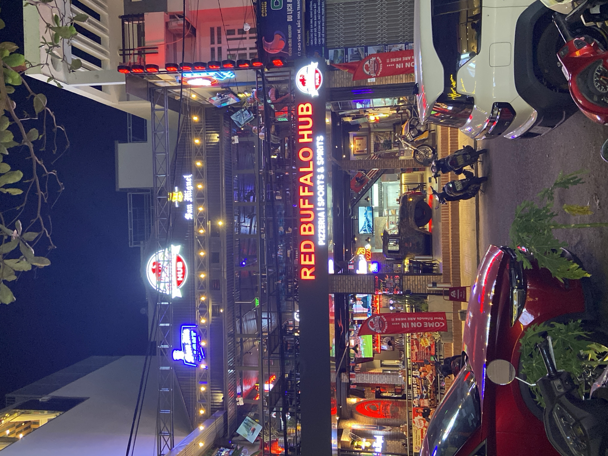 The image captures a vibrant street scene at night in Bac Nha Trang Ward, Vietnam. A multi-story building with a prominent neon sign reading "RED BUFFALO HUB" dominates the view. The establishment appears to be a pizzeria, sports bar, and spirits venue, indicated by the text "PIZZERIA | SPORTS & SPIRITS" below the main sign.
The building's facade is illuminated by numerous neon lights and string lights, creating a lively atmosphere. Several levels of the building are visible, with balconies and tables where patrons are seated, some of whom are blurred due to motion. Various signs and posters are displayed, including one in Vietnamese that translates to "COME ON IN.... Your friends are here!!" and another that mentions "WORLD CUP LIVE & LOUD".
In the foreground, two vehicles are parked on the street: a red car and a white SUV. Several motorcycles are also present. The street is paved, and there are some plants and trees lining the sidewalk. The overall impression is of a bustling urban nightlife scene in Vietnam.