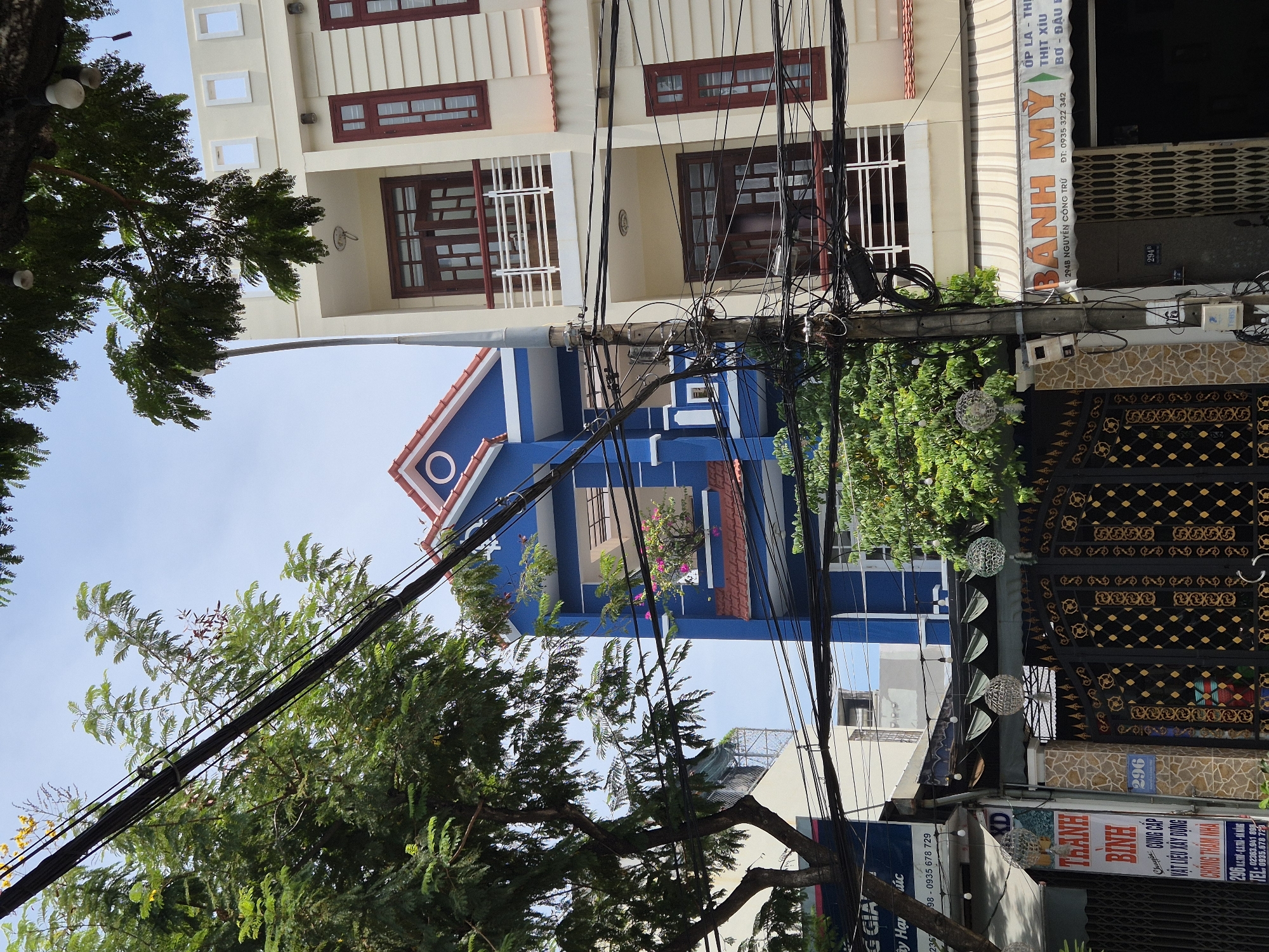 The image shows a street scene in Đà Nẵng, Vietnam. It features a narrow, multi-story blue building with a red roof, a cream-colored building with red-trimmed windows, and a building with a red awning that reads "BÁNH MỲ" (Vietnamese for baguette sandwich). There are numerous electrical wires and a utility pole dominating the foreground. Trees with green foliage are visible on the left and right sides of the frame. Decorative spherical lights hang from the trees and near the buildings. A black metal gate with gold embellishments is visible on the left. Signs in Vietnamese are present on the buildings, indicating businesses such as a bakery and a construction supply company. The sky is visible and appears to be bright and clear.