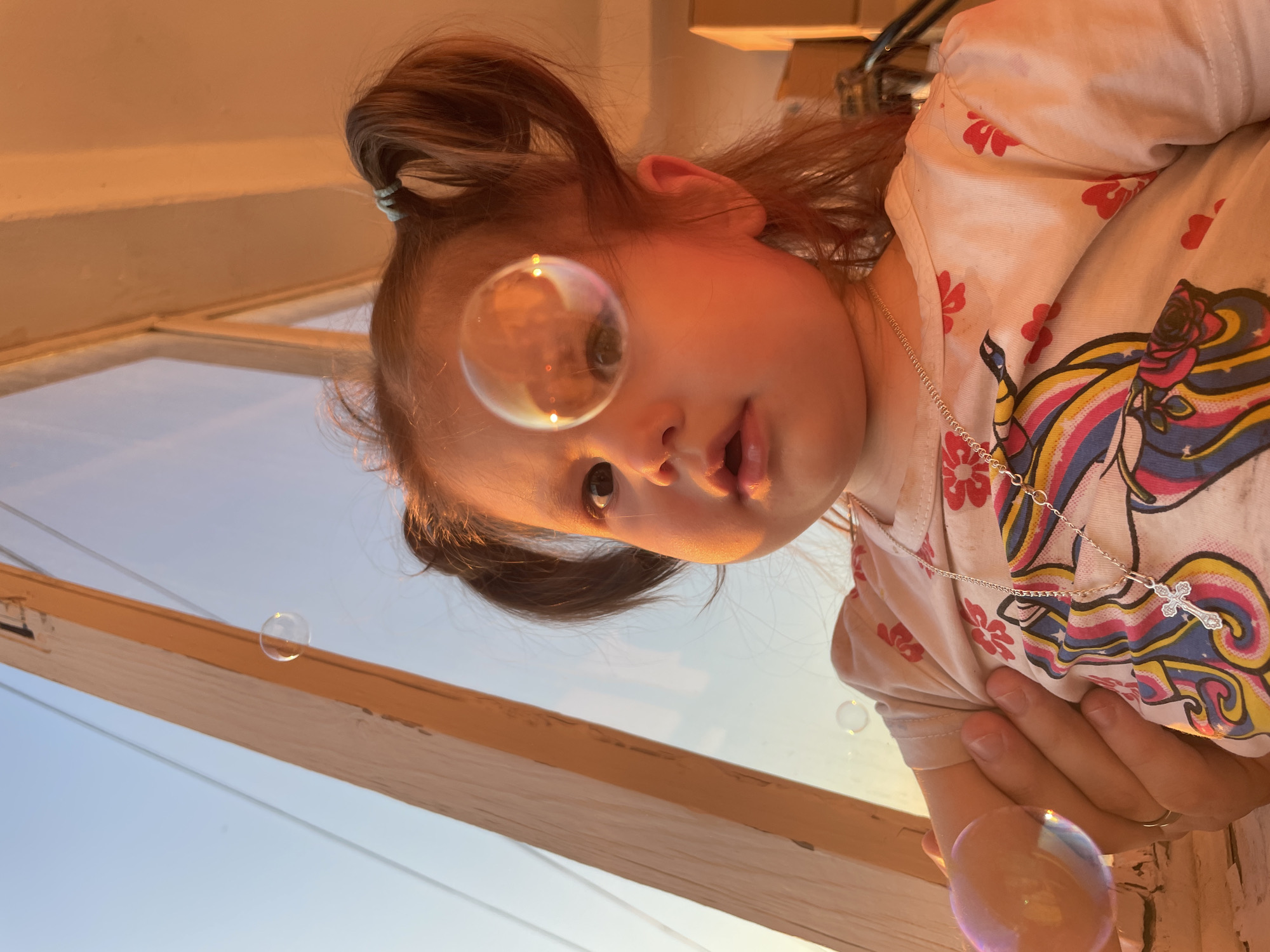 This image depicts a low-angle, close-up portrait of a young child, likely a toddler, being held by an adult during sunset or sunrise. The child has light brown hair styled into two pigtails, secured with light blue hair ties. The face is round with large dark eyes and slightly parted lips. A silver chain necklace with a small, clear-stoned cross pendant hangs around the child's neck. The child wears a light-colored t-shirt adorned with a colorful graphic design featuring a unicorn or similar mythical creature with a rainbow mane and floral elements, alongside scattered smaller red floral or heart patterns.
An adult's left hand, wearing a ring on the ring finger, is visible supporting the child's left arm and torso. Several translucent, iridescent soap bubbles are present in the scene. One large bubble floats prominently in front of the child's face, reflecting the warm light and surrounding environment, and partially obscuring the child's left eye. Another smaller bubble is visible in the lower left, and a third, even smaller bubble is in the upper left background, beyond the window.
The background features an interior setting bathed in warm, golden light characteristic of late afternoon or early evening. A light-colored wooden vertical window frame or support beam is prominent on the left. Through the window, a clear sky is visible, transitioning from a pale blue at the top to a warm orange-yellow nearer the horizon. Portions of an interior wall, also illuminated by the warm light, are visible in the upper right. This scene is situated in the city of Izhevsk, Russia.
