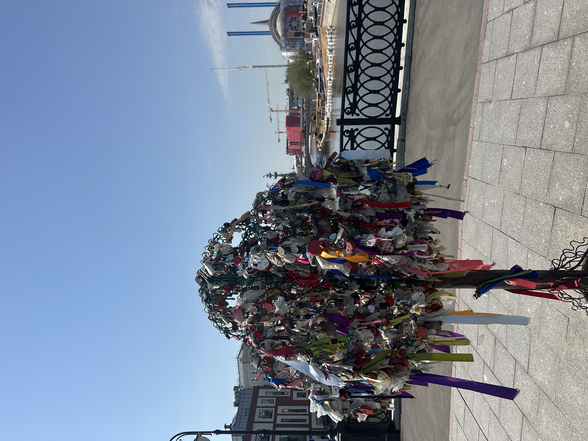 This image shows a love lock tree on Luzhkov Bridge in Moscow, Russia. The tree is covered with locks and ribbons left by couples as a symbol of their love. The Moskva River and a construction site with cranes are visible in the background. The sky is clear and blue. The tree is located on a paved walkway. A black metal railing runs along the riverbank.