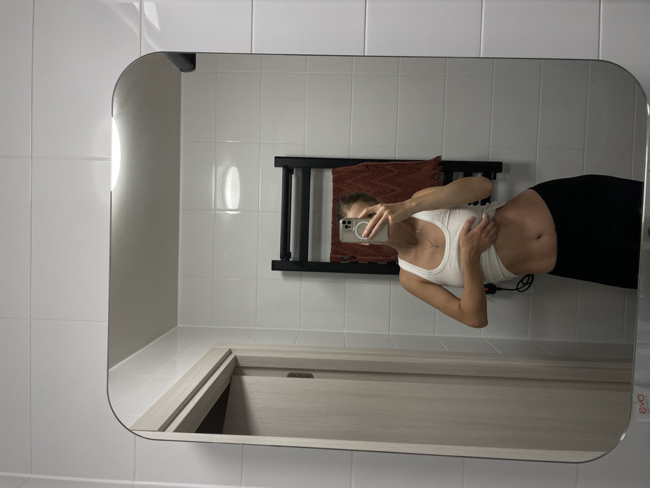 A mirror reflects a bathroom scene with white tiled walls and a light wooden vanity with a drawer. A black heated towel rack holds a dark orange towel. Reflected in the mirror is a person, presumably taking the picture, wearing a white tank top and black leggings. Their midriff is visible. They are holding a phone in front of their face, obscuring it. A silver necklace is visible.