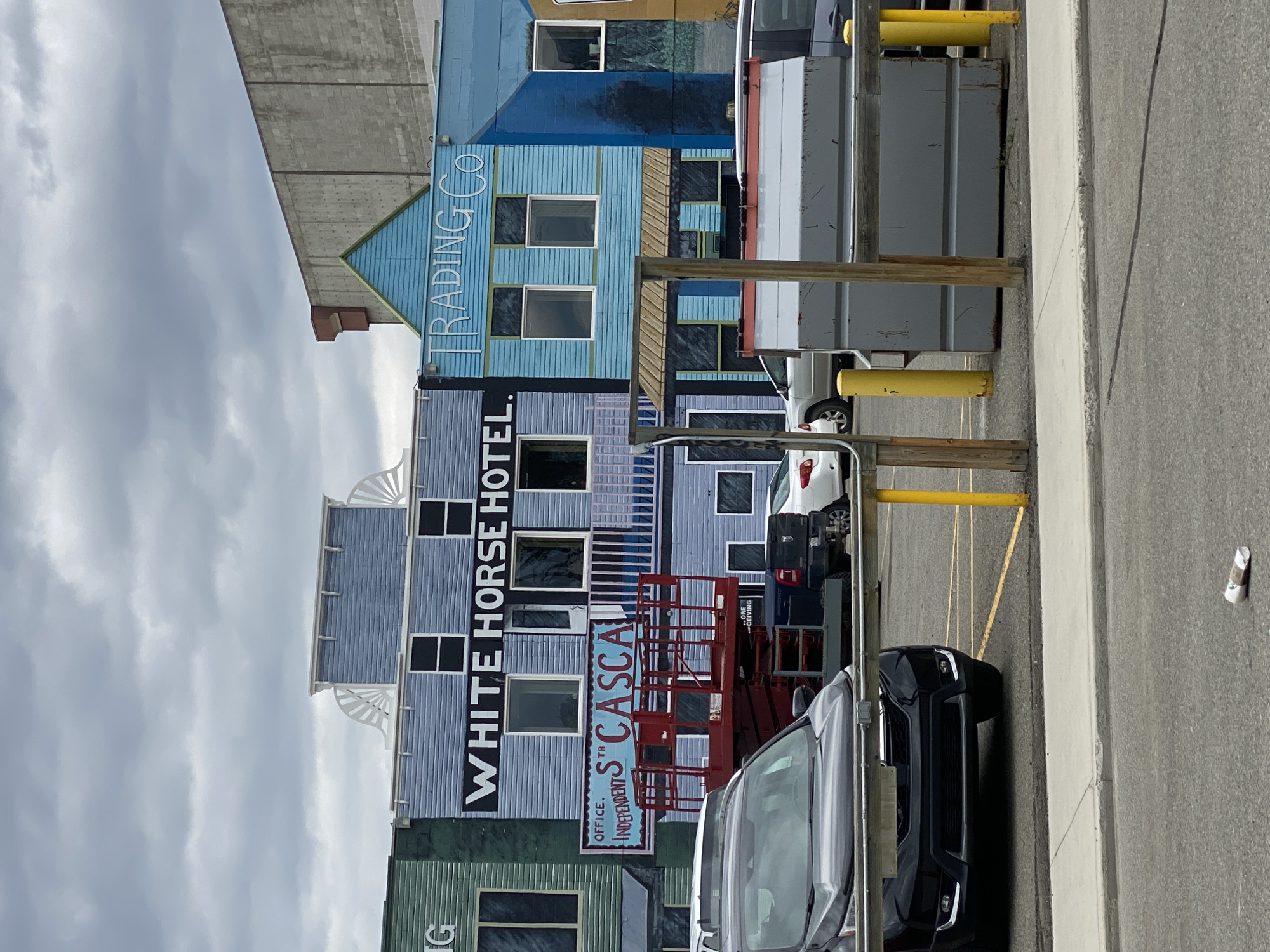 The image shows a street scene in Whitehorse, Canada, featuring the facades of several buildings painted in a colorful, Western-style mural. The central building is labeled "WHITE HORSE HOTEL" in large black letters. To its left, a green building with the partial word "IG" is visible, and to its right, a light blue building is labeled "TRADING CO". Below the "WHITE HORSE HOTEL" sign, a smaller sign reads "OFFICE. INDEPENDENTS ST CASCA". A red hydraulic scissor lift is positioned in front of the "WHITE HORSE HOTEL", with its platform partially raised. Several cars are parked in a lot in front of the buildings. The sky is overcast with clouds. In the foreground, there is a sidewalk and a paved area with yellow bollards and a large grey dumpster. A crumpled paper cup is on the ground in the lower right corner of the image.
