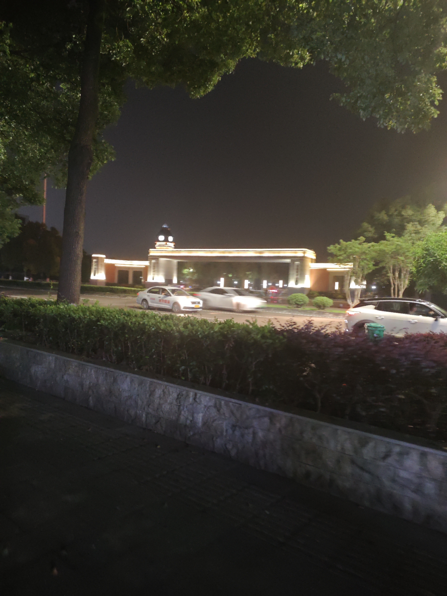 The image depicts an outdoor scene at night, likely in the Chongqing High-tech Zone, China. In the background, a building with illuminated archways and a clock tower is visible. Several cars are driving on a road in front of the building. One white car, possibly a taxi, has red text on its side. Green bushes and trees line the road, and a stone wall separates the road from the foreground. The overall atmosphere is dimly lit due to the nighttime setting and the presence of artificial lighting from the building and streetlights.