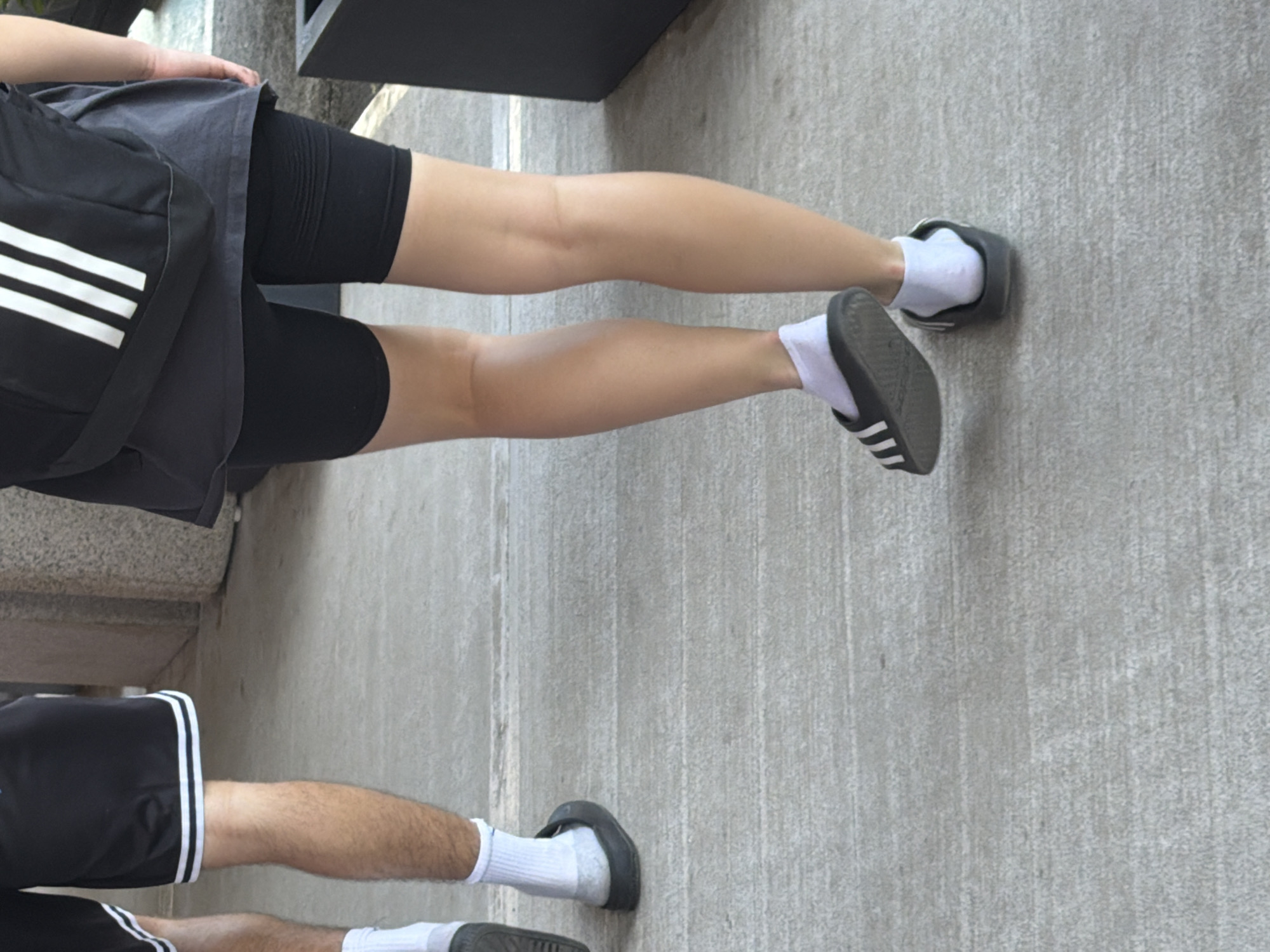 An outdoor, eye-level shot captures the lower halves of two individuals standing or walking on a light gray concrete surface. The primary subject, positioned centrally, wears a dark grey top over black biker shorts that feature a ribbed texture on the upper thigh. A black shoulder bag with three distinct white stripes, indicative of the Adidas brand, hangs across their side. Their bare legs are visible, extending down to white ankle socks and black Adidas slides with white stripe detailing on the straps.
To the lower left, the legs of a second person are partially visible. This individual wears black shorts with white trim along the hem. Similar to the first person, they are also wearing white ankle socks and black Adidas slides. Their legs appear hairier than the first subject's.
The background consists of a uniform concrete ground and a dark, indistinct structure or wall in the upper right. The bright, even lighting suggests it is daytime. The overall scene conveys a casual atmosphere, with both individuals sporting active or leisure wear, including prominent Adidas branding on their accessories and footwear.