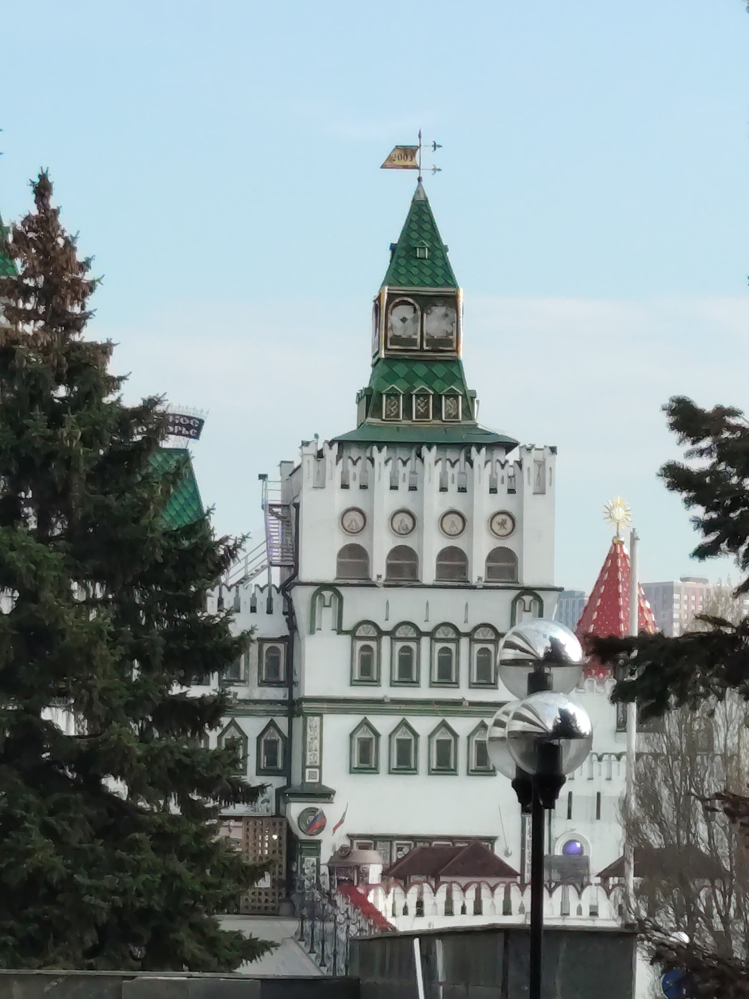 This image presents a picturesque view of an ornate, white tower with a green, multi-tiered conical roof, characteristic of traditional Russian "fairy tale" architecture. The structure is partially framed by dense, dark green coniferous trees on both sides, suggesting a park-like or expansive setting.
The central tower is highly detailed, featuring a weathervane atop its spire that clearly displays the year "2003". Below the spire, the green roof transitions into a white battlemented section adorned with several small, round arched windows, and further down, rows of intricately framed arched and rectangular windows. The white walls are accented with green trim, enhancing the distinctive architectural style.
To the left of the main tower, another building with a green roof is partially visible behind a prominent evergreen tree. On its roof, a sign in Cyrillic can be discerned, reading "1500 ДОБРЫ...". To the right, peeking from behind the main tower, is a striking red conical roof crowned with a golden, sun-like finial. In the foreground, a modern lamppost with two reflective spherical lights stands out.
The scene is captured on a clear day, indicated by the light blue sky, and appears tranquil with no people visible. The architectural style, combined with the Moscow context, strongly suggests this is the Izmailovsky Kremlin, a cultural and entertainment complex. The overall impression is one of historical charm and architectural splendor.
