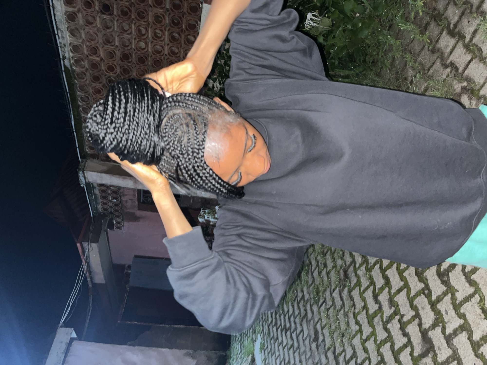 A young Black woman with braided hair pulled up in a bun is standing outdoors at night, adjusting her hair. She is wearing a dark gray long-sleeved sweatshirt and green shorts. The background shows a building with a textured wall, some foliage, and a paved ground with some grass growing between the pavers. The image appears to be taken in a residential area.