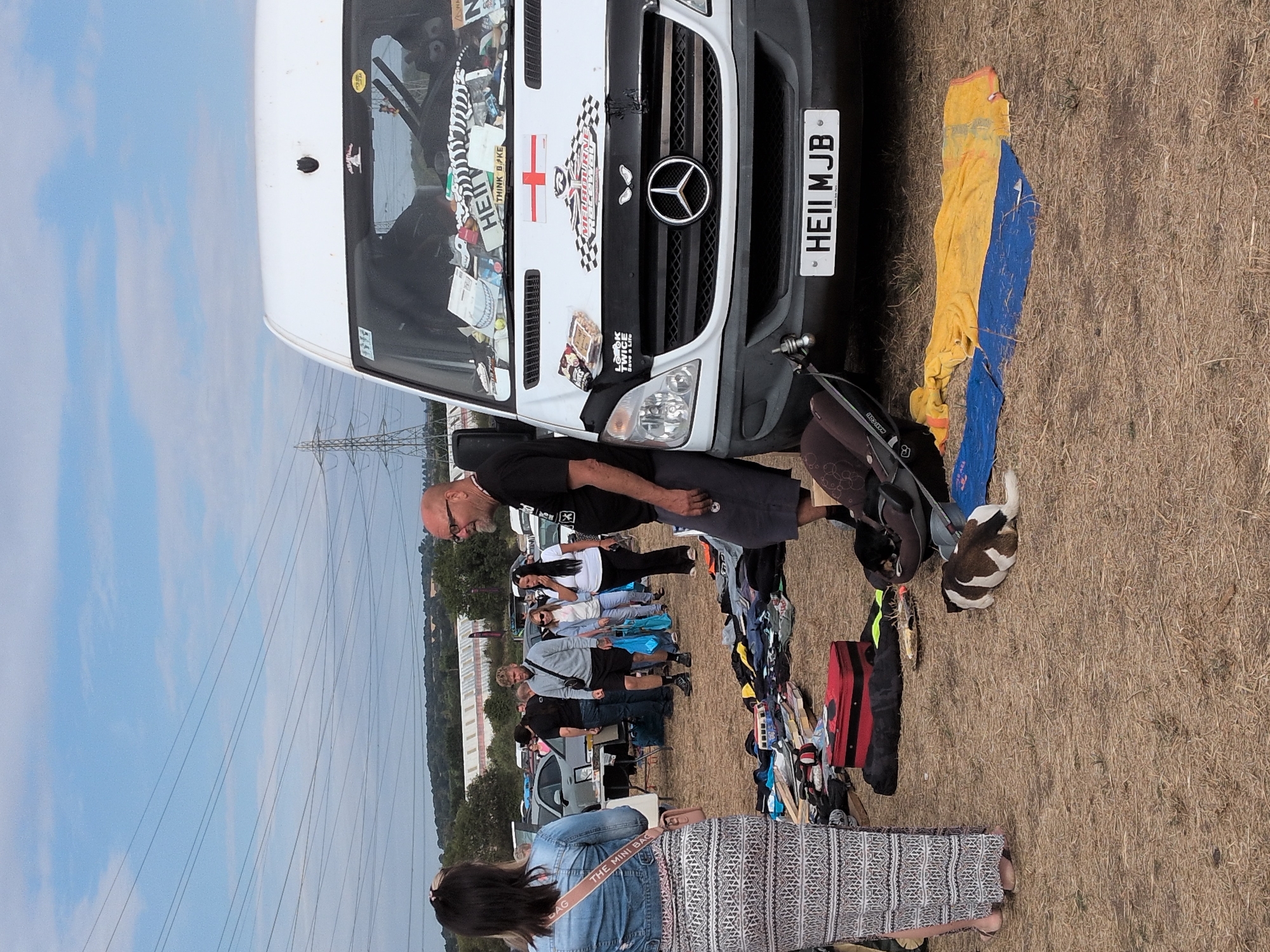 This image depicts an outdoor market scene, likely a car boot sale, under a partly cloudy sky. In the foreground, a woman wearing a long patterned skirt and a denim jacket stands facing away from the camera. To her right, a man with glasses and a black t-shirt stands near the front of a white Mercedes-Benz Sprinter van. The van is adorned with various stickers, including a St. George's Cross flag and checkered racing decals. A license plate reading "HEII MJB" is affixed to the front. Scattered on the ground around the van are various items, including clothing, a red suitcase, and a car seat. Two dogs are visible on the ground; one is lying down, and the other appears to be a puppy. In the background, several other individuals are browsing stalls and vehicles, with more cars and a large building visible in the distance. Power lines crisscross the sky. The overall setting suggests a relaxed, informal event in a rural or semi-rural location. The presence of power pylons and a distant industrial-looking building suggests the location is near a developed area within South Staffordshire, United Kingdom.