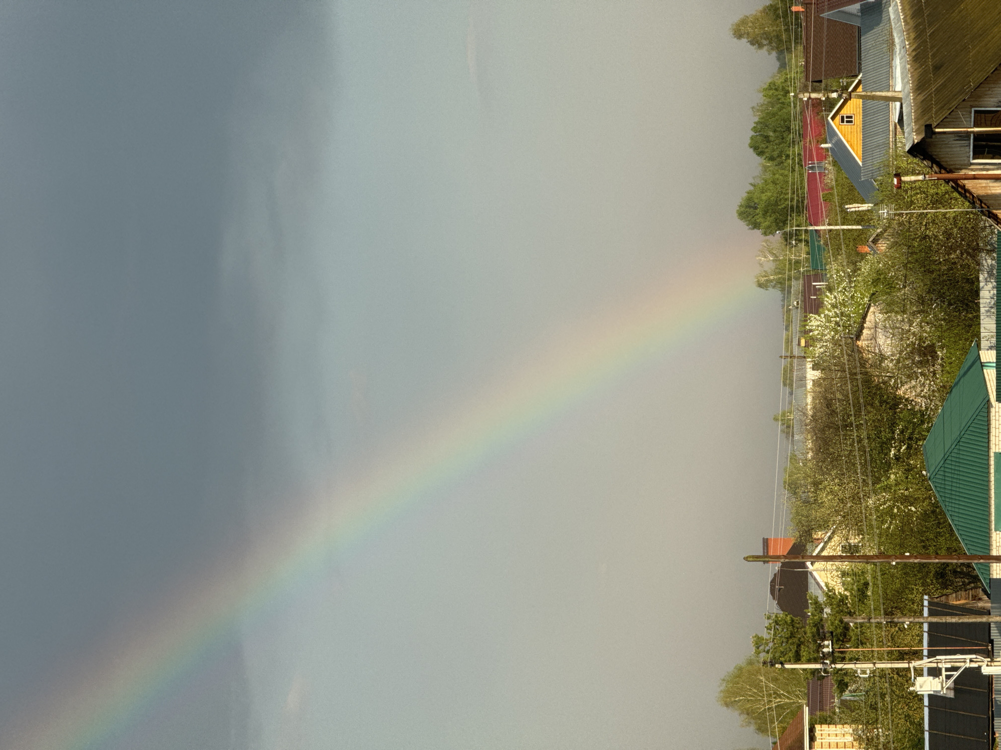 The image shows a rainbow arcing across the sky above Bol'shoye Kozino, Russia. The left side of the frame is dominated by a dark, stormy sky. The rainbow's colors are visible, transitioning from violet to red. On the right, the rainbow terminates over residential structures with varied roof colors, including red, yellow, and green. Utility poles and wires are visible amongst blooming trees.
