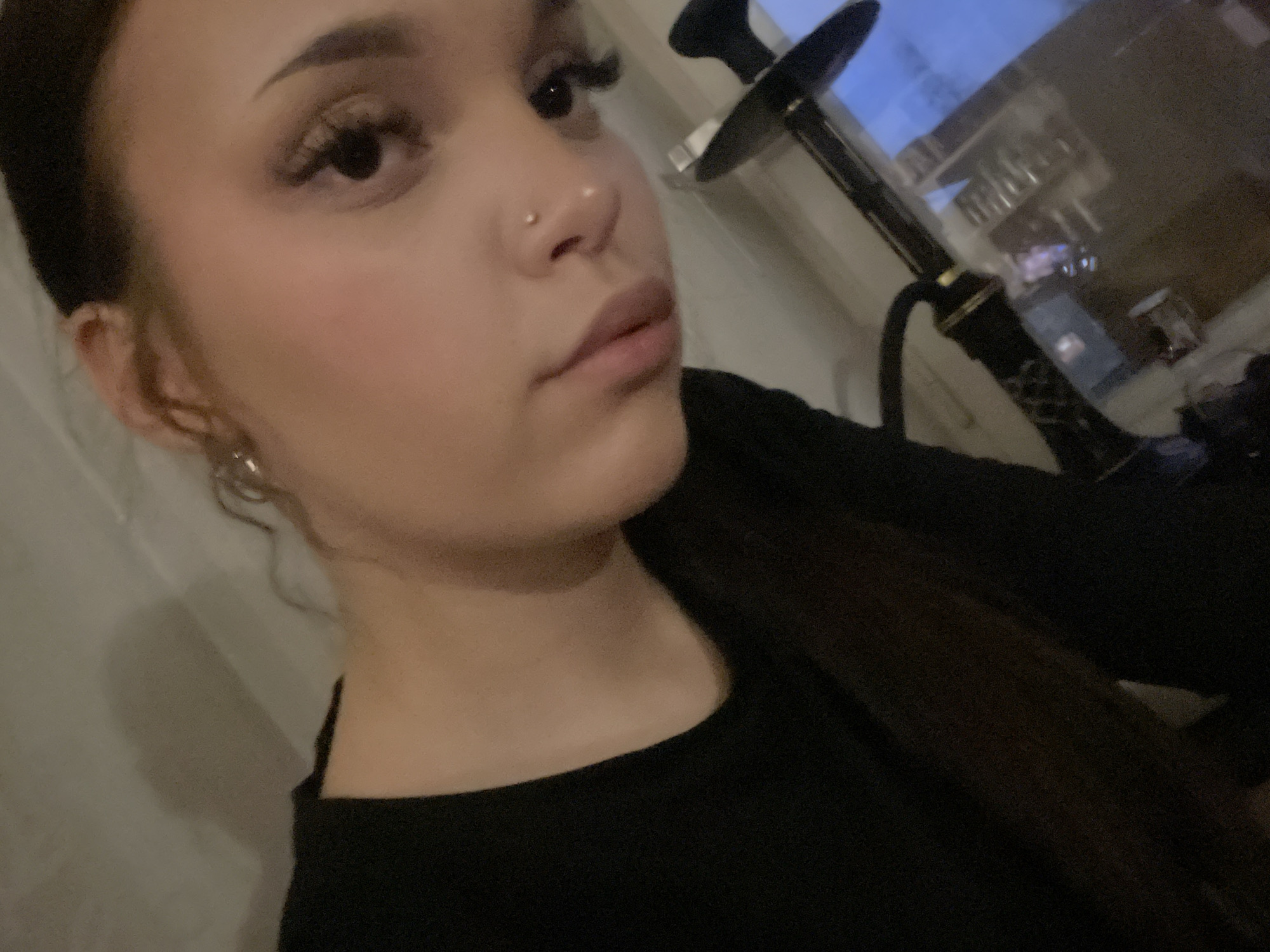 The image displays a young woman indoors, captured from a close-up, slightly upward angle, focusing on the right side of her face and upper body. Her dark hair is pulled back or styled to the side, revealing a metallic earring on her right ear. She wears makeup, including prominent eyelashes, eyeshadow, and blush, and has a small stud piercing on her right nostril. Her expression is neutral, with her gaze directed slightly away from the camera. She is dressed in a dark, likely black, top.
In the blurred background, a shisha or hookah pipe is partially visible to the right of the woman, positioned near a window. The window's reflection suggests an urban environment at dusk or night, with faint lights and indistinct structures, indicating an evening setting. The overall ambiance is dimly lit and relaxed. There is no visible text in the image. Although the context of Saint Petersburg, Russia is provided, no explicit visual cues within the photograph confirm this specific location.