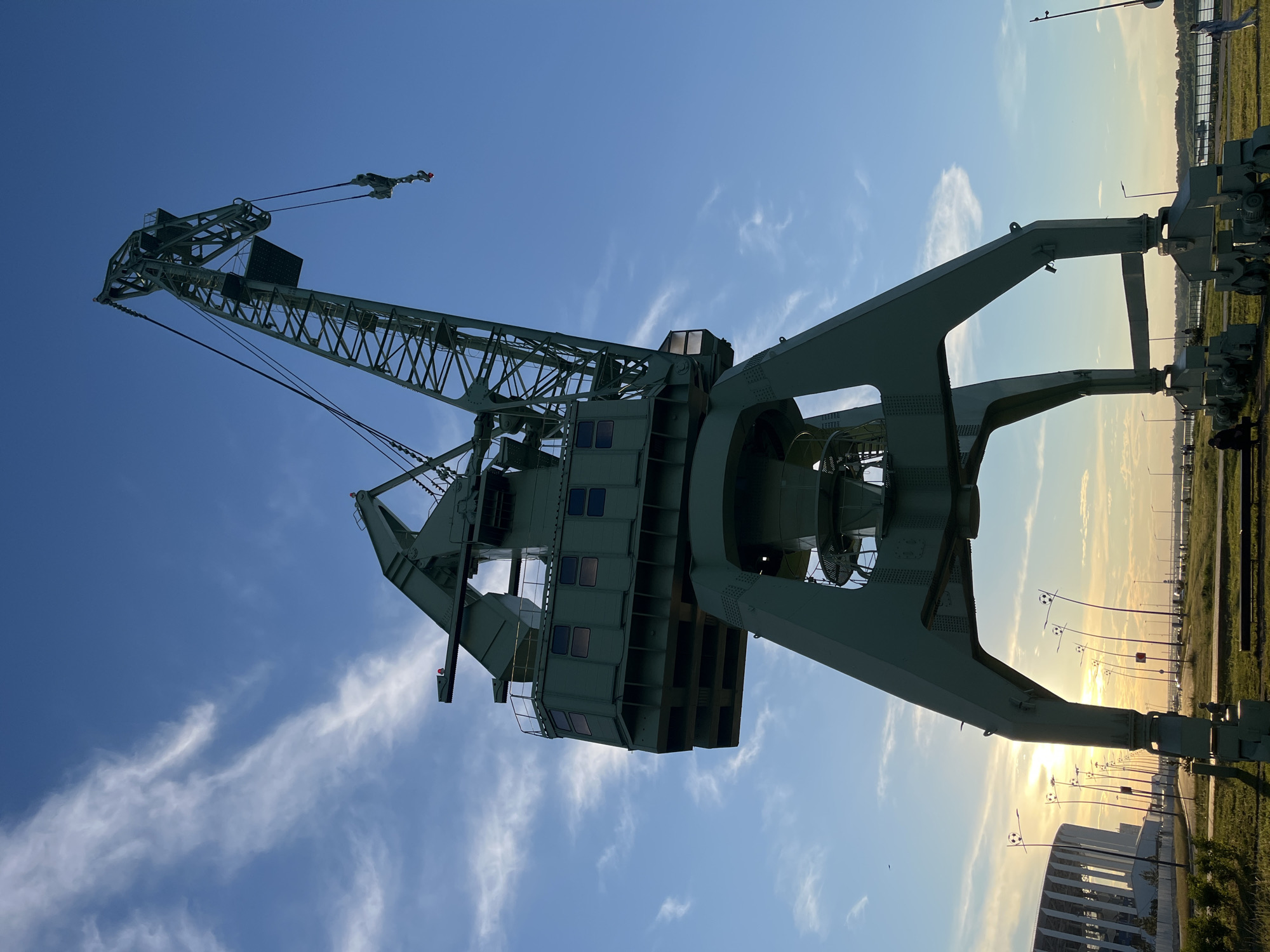 This image captures a large, green gantry crane against a bright blue sky with wispy clouds. The crane dominates the frame, its intricate metal structure visible as it extends upwards and outwards. The sun is setting, casting a warm glow on the landscape visible in the distance, which includes grassy fields, streetlights, and modern buildings. The scene appears to be an industrial or port area, possibly in Nizhny Novgorod, Russia, given the location context. The low angle of the sun suggests it is late afternoon or early evening. There are no people prominently visible in the shot, but a few figures can be faintly seen in the distance near the buildings. The overall mood is one of industrial grandeur and the calm of a day's end.