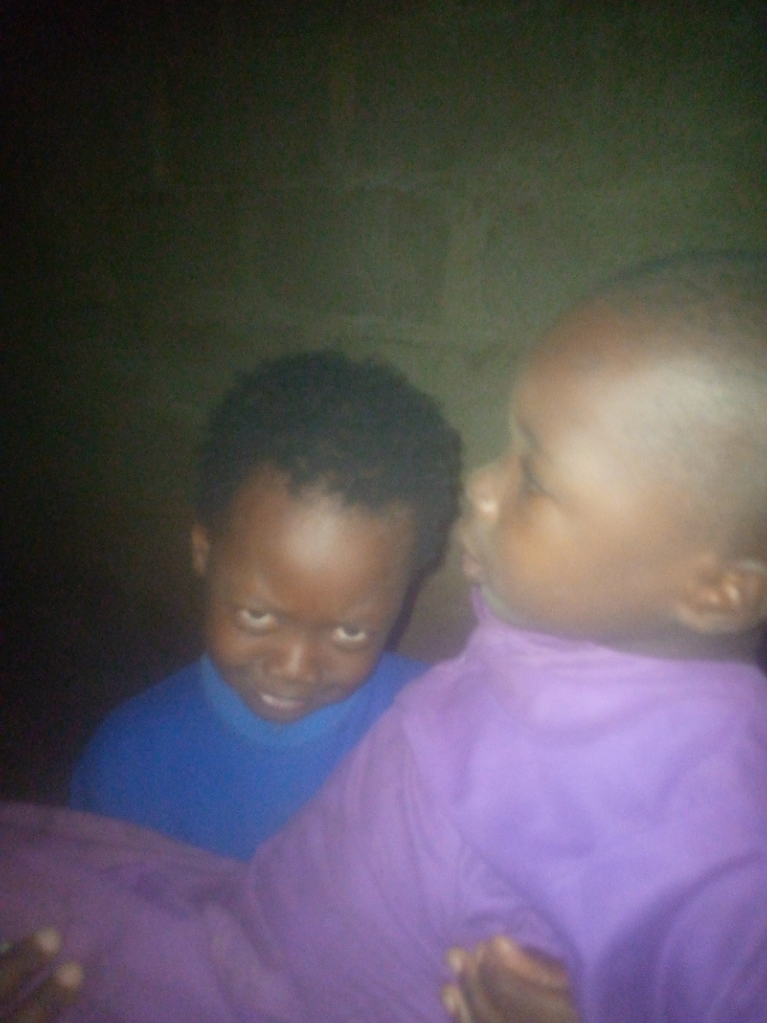 The image captures two young children indoors, photographed in low light conditions. In the foreground, on the right side, is a child with dark skin, depicted in profile, facing towards the right edge of the frame. This child is wearing a purple long-sleeved top and appears calm, though their full expression is not clearly visible.
Behind and slightly to the left of the foreground child, another child with dark skin looks directly at the viewer. This child is wearing a blue top and has an intense, somewhat serious expression with slightly raised eyebrows, suggesting curiosity or a playful gaze. Both children appear to be toddlers or very young.
The background is uniformly dark and indistinct, offering no specific location cues beyond an indoor setting, possibly a wall. The low, dim lighting suggests the photograph was taken in the evening, at night, or in a poorly lit room, contributing to the somewhat grainy and blurry quality of the image. No text is visible in the frame.
