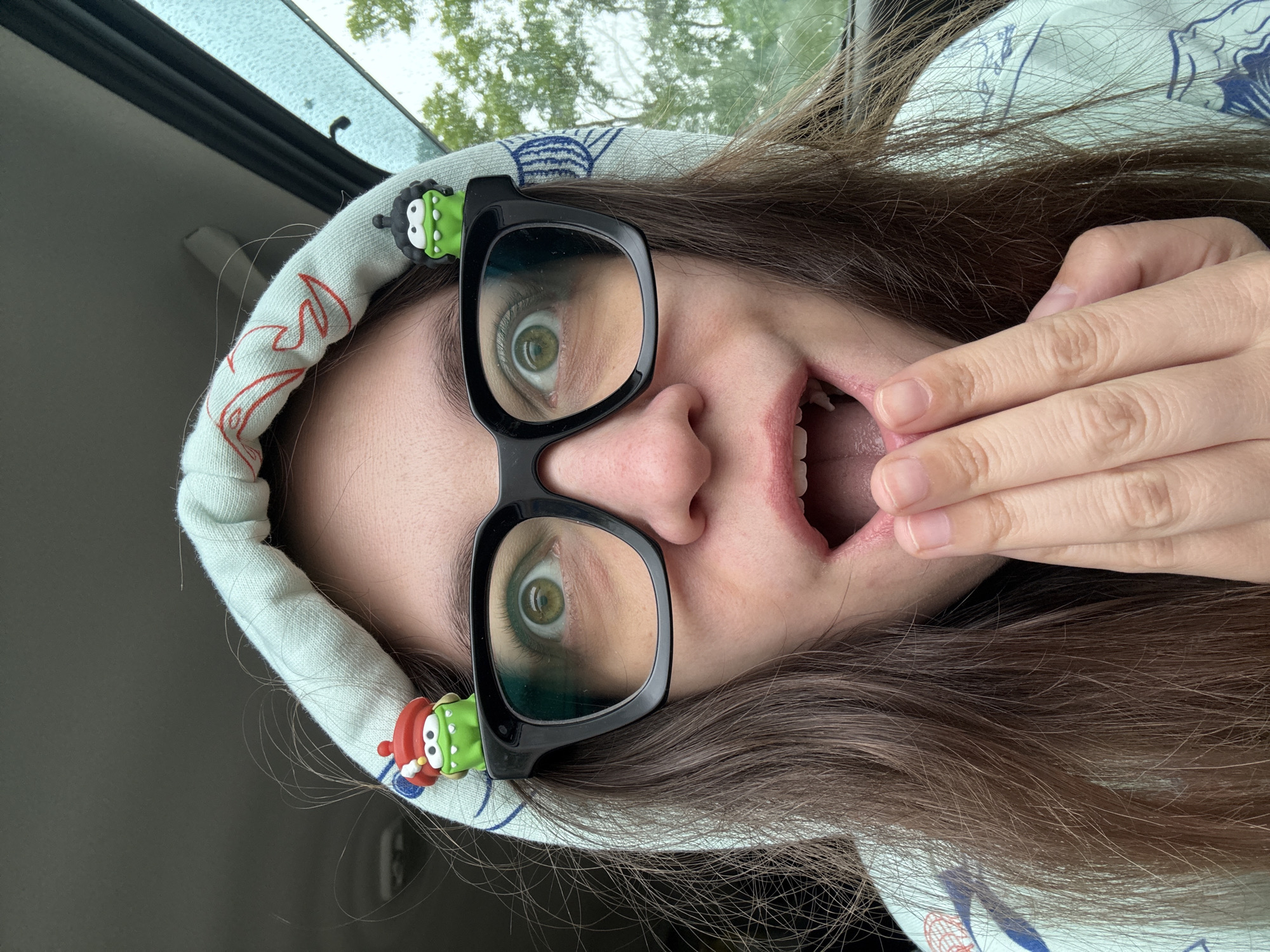 A young woman is shown from a skewed, close-up perspective. She is wearing black-framed glasses with cartoonish green monster decorations attached to the temples. The woman has brown hair and is wearing a light blue hooded sweatshirt with a pattern on the hood. Her mouth is open, and her hand is near her mouth as if she is either speaking or reacting to something. The background shows part of a car interior and a glimpse of trees and sky, suggesting she is inside a vehicle. The location is not discernible from the image and cannot be pinpointed as Фоки, Russia.