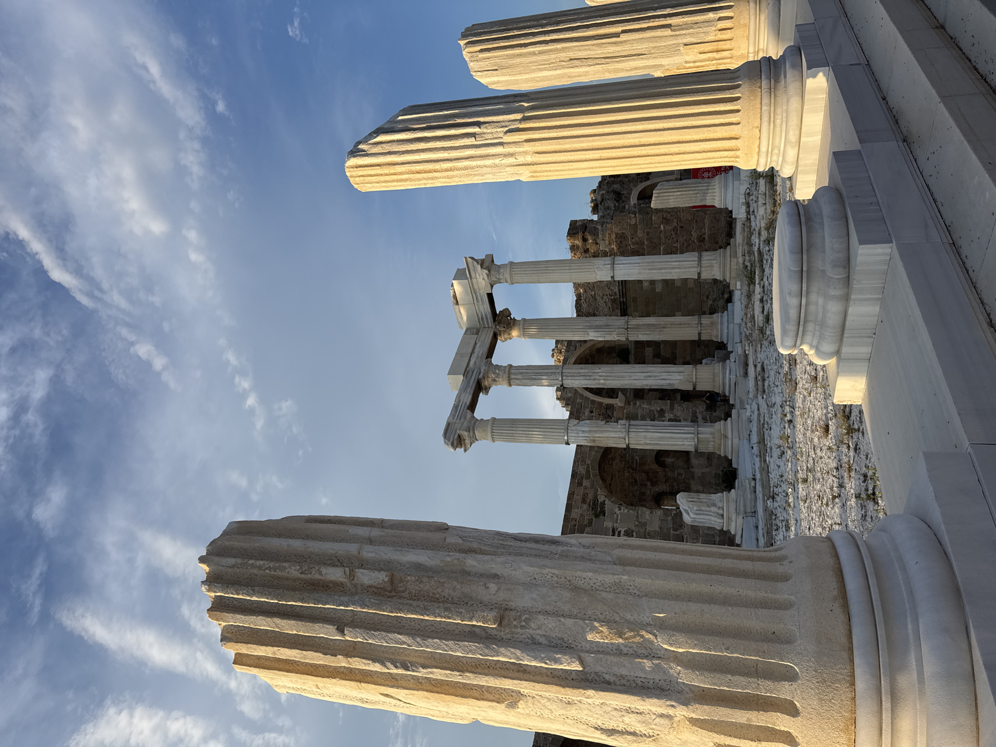 The image shows the ruins of the Temple of Apollo in Side, Turkey. The foreground features large, fluted marble columns, with one prominent column on the left casting a shadow. Behind these, more columns form an archway and a colonnade. The ruins consist of stone walls and arches. The ground is covered with gravel and sparse vegetation. The sky above is a clear blue with wispy white clouds, illuminated by what appears to be late afternoon or early morning sunlight, casting a warm glow on the ancient structures. Two indistinct figures can be seen in the background near the ruins.
