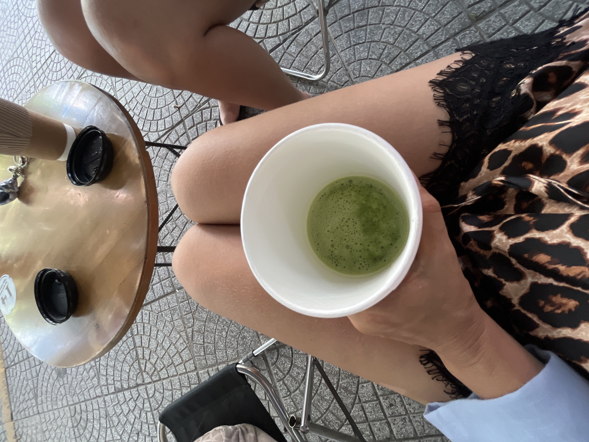 The image depicts a first-person, downward-facing view of a cafe scene in Đà Nẵng, Vietnam. In the immediate foreground, the bare thighs of an individual are visible, clad in a leopard print skirt with black lace trim on the right side, and a light blue long-sleeved top on the left. The individual's hands are clasped around a white disposable cup containing a green, frothy beverage, consistent with matcha.
Adjacent to the primary subject's legs, on the right side of the frame, the bare legs and foot of a second individual are partially discernible. Situated between the two individuals is a small, round wooden table with a dark metal base. Resting on the table are two black plastic lids, a light brown ribbed disposable coffee cup, and a metallic keychain with keys. A white circular sticker featuring a "1+" symbol is affixed to the tabletop.
The ground surface is covered with grey concrete tiles displaying a repeating geometric pattern of interconnected squares and partial circles. A portion of a metal chair frame with black fabric is visible on the far left, partially obscured.
