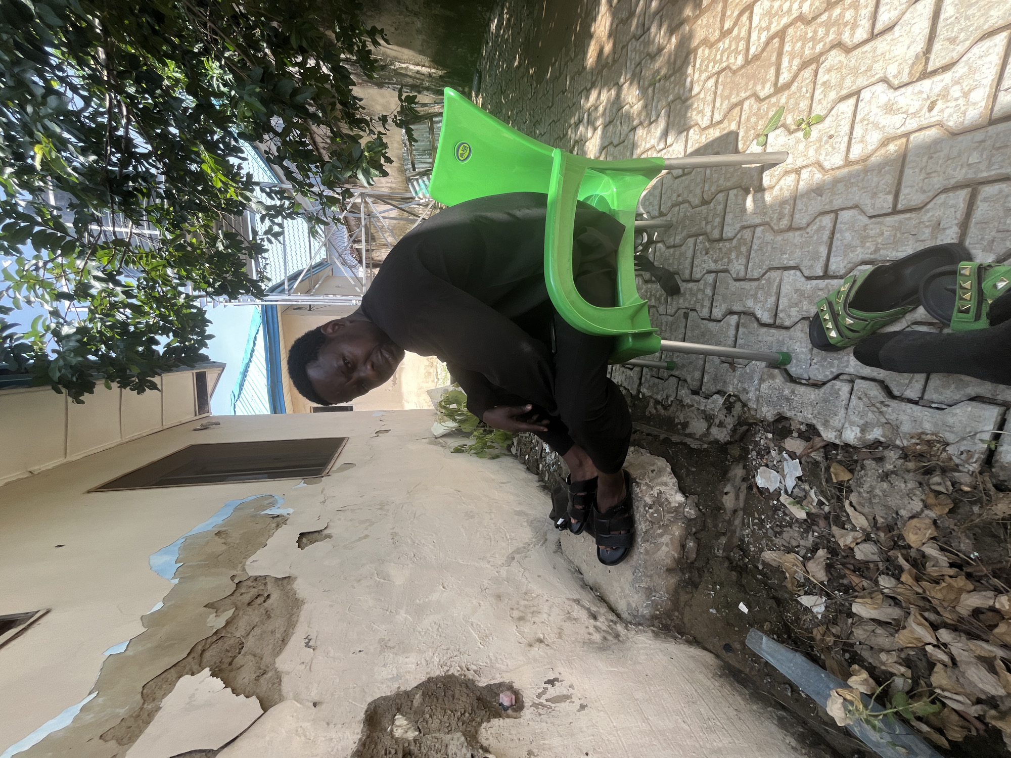 A man sits on a green plastic chair in front of a weathered building in Galadimawa Village, Nigeria. The building's exterior wall is damaged, with peeling paint and exposed concrete. The ground is paved with interlocking concrete blocks, and debris including dry leaves and broken pieces of material are scattered near the wall. The man is wearing black clothing and black sandals. In the foreground, a pair of green sandals with metallic stud details are placed on the paved ground, suggesting another person was present or is nearby. Sunlight casts shadows on the ground, indicating it is daytime. Trees with green foliage are visible in the background, partially obscuring some of the surrounding structures.