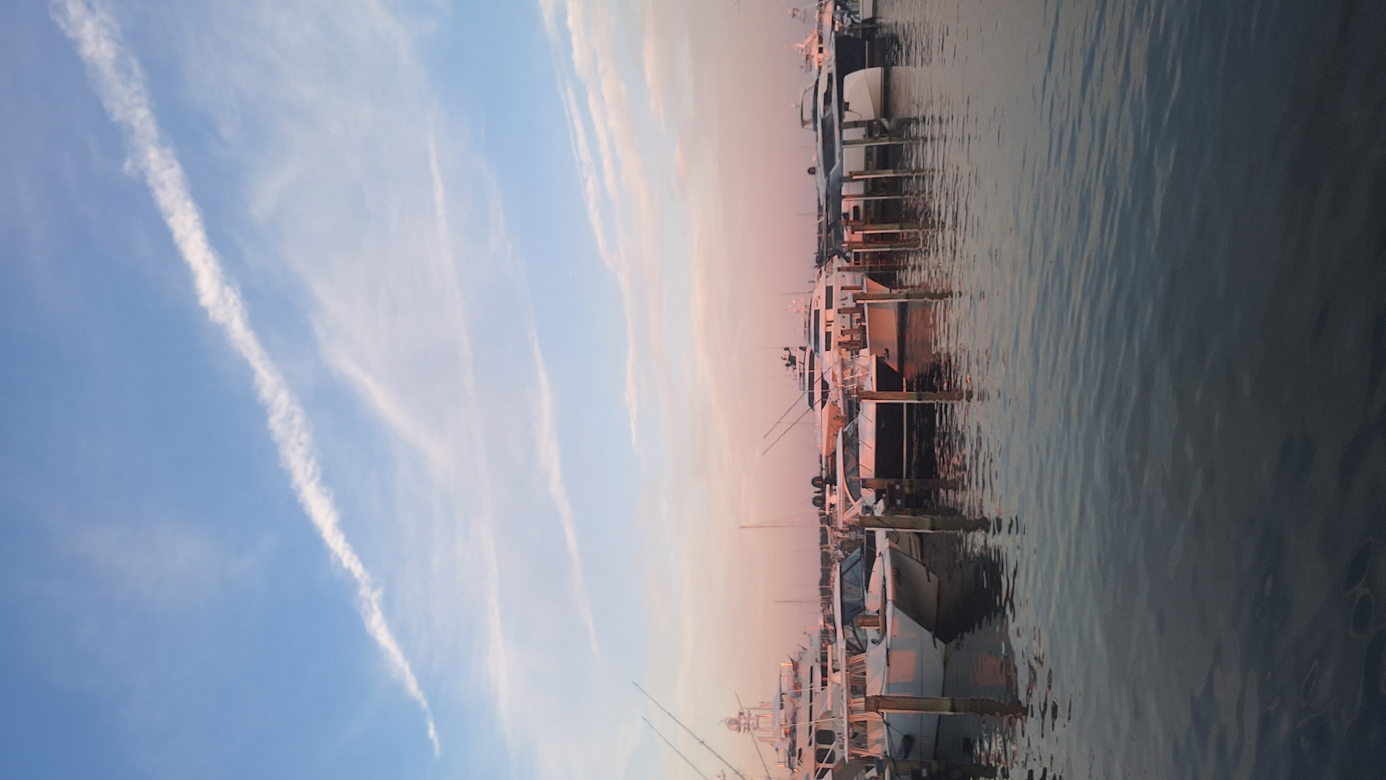 The image depicts a harbor filled with several docked boats of varying sizes, possibly yachts and smaller motorboats, illuminated by the soft light of a sunset. The sky above is a blend of pale blue and soft pink, streaked with wispy white clouds, suggesting the golden hour. The water in the foreground is calm and reflective, mirroring the colors of the sky and the boats. The boats are tightly packed in the marina, with some showing fishing rods extending upwards. The scene suggests a peaceful evening in a coastal city. The location is identified as Nantucket, United States of America.