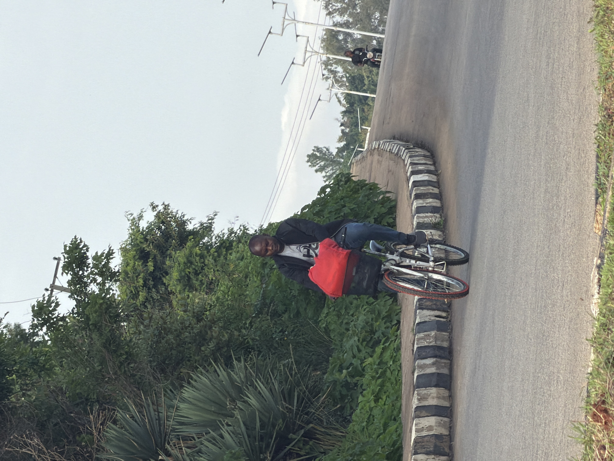A man is riding a bicycle on a paved road that curves uphill. He is wearing a black jacket, a grey t-shirt, and blue jeans. A bright red bag is attached to the front of his bicycle. The road is lined with a decorative concrete barrier painted with alternating black and white stripes. Lush green vegetation, including large spiky plants and trees, grows on the left side of the road. In the background, there are streetlights and utility poles, as well as another person on a motorcycle further up the road. The sky is overcast and light in color. The scene appears to be on an uphill road in Bukuru, Nigeria.