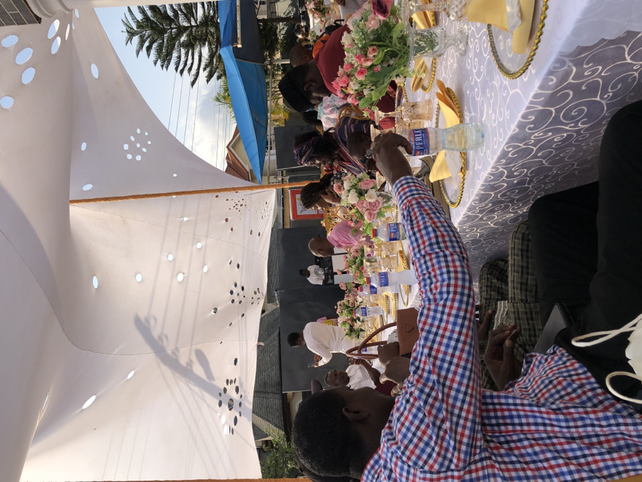 The scene depicts an outdoor social gathering or event in the city of Unknown, Ghana, occurring during daylight hours under a large, white fabric canopy. The canopy features various circular and decorative cutouts, casting fragmented shadows. Multiple individuals, primarily adults, are seated at long tables covered with white tablecloths displaying a raised, swirling pattern. Each place setting includes a gold-rimmed charger plate, a yellow napkin, and clear glassware. Floral centerpieces, composed of pink, white, and green flowers, are distributed along the tables, accompanied by "Perla" brand bottled water.
In the left foreground, a man in a red, white, and blue plaid shirt is seated, extending his arm towards a water bottle on the table. A brown handbag rests on the table near him. In the mid-ground, a man in a white long-sleeved shirt stands, holding a mobile phone in his right hand, facing towards the right side of the image. Several other guests, including a woman wearing a patterned headwrap and a man in a pink collared shirt, are seated at tables in the mid-ground. Towards the left background, a man stands behind what appears to be a DJ setup. The immediate background shows a building with a dark-tiled roof on the left, and a tall evergreen tree, a blue tarp structure, and a white architectural pillar on the right. Black paneling is visible behind the standing man in white.